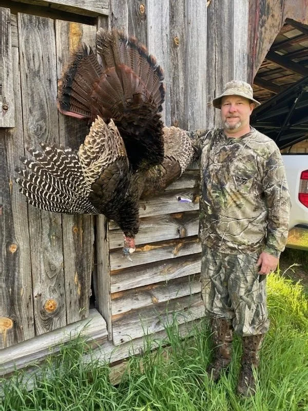 A man in camouflage clothing and a hat stands next to a wooden shed, smiling, with a large turkey mounted on the shed's wall.