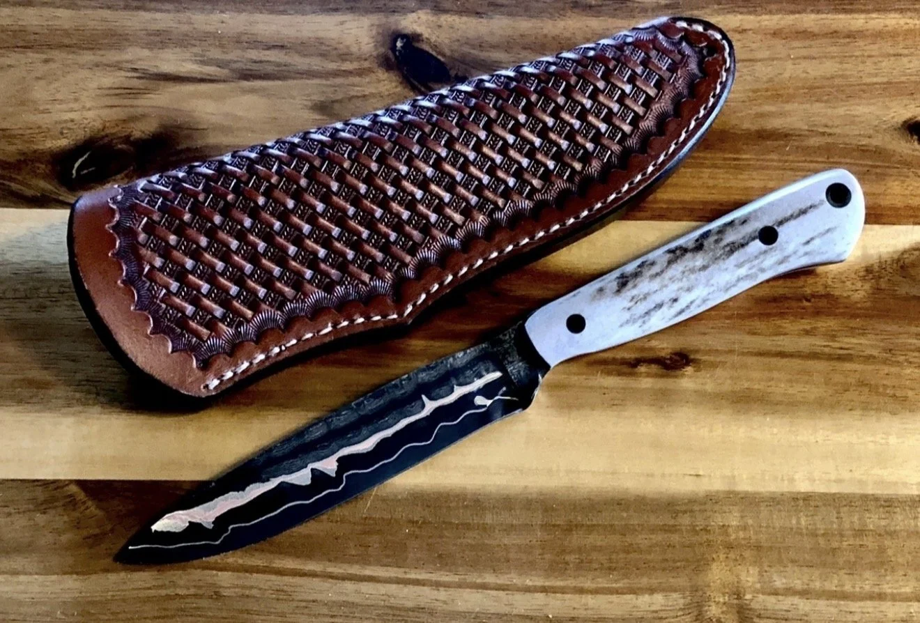 A hunting or camping knife with a black and silver patterned blade and a white wooden handle, resting on a wooden surface next to a leather sheath with a woven design.