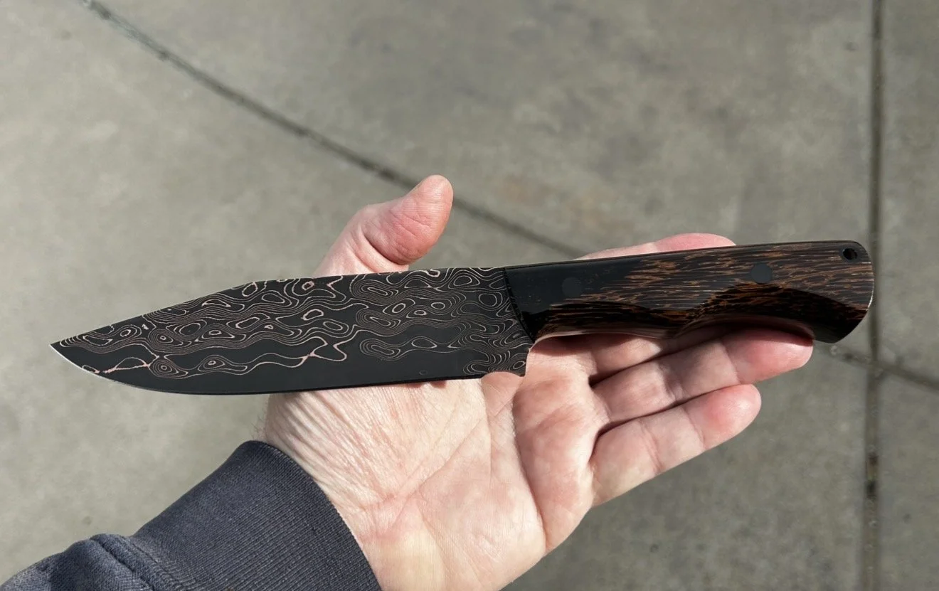A hand holding a black bent knife with a Damascus pattern blade and a wood grain handle.