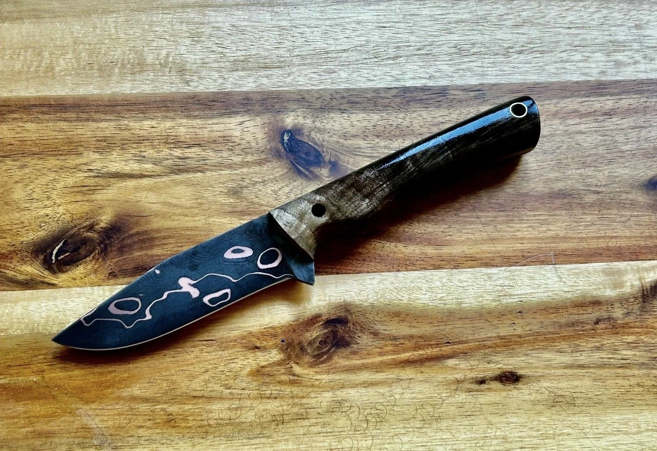 A black folding pocket knife with a decorated handle, placed on a wooden surface.