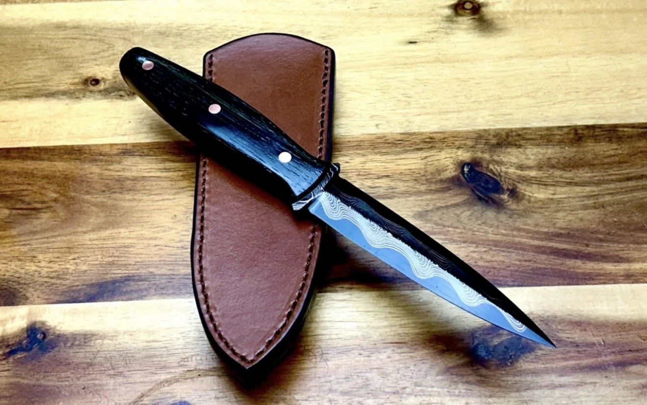 A folding pocket knife with a black handle and a Damascus steel blade, resting on a brown leather sheath on a wooden surface.