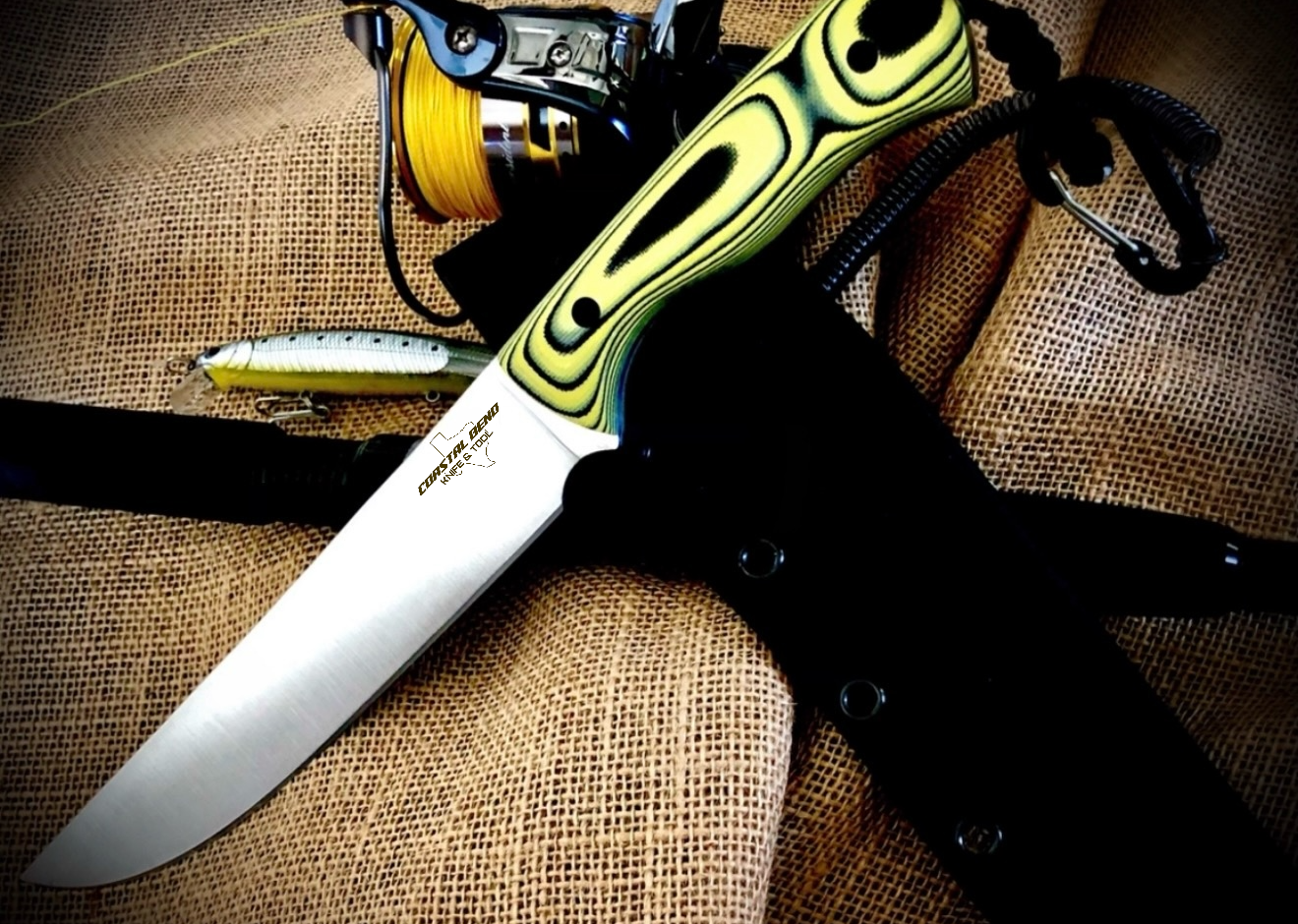 A large fixed-blade knife with a yellow and green handle resting on a textured brown surface, with a fishing reel, fishing line, a fishing lure, and a black sheath nearby.