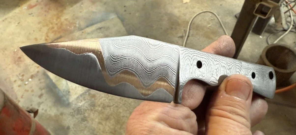 Close-up of a handcrafted knife with a Damascus steel blade and a plain steel handle, held by a person's hand.