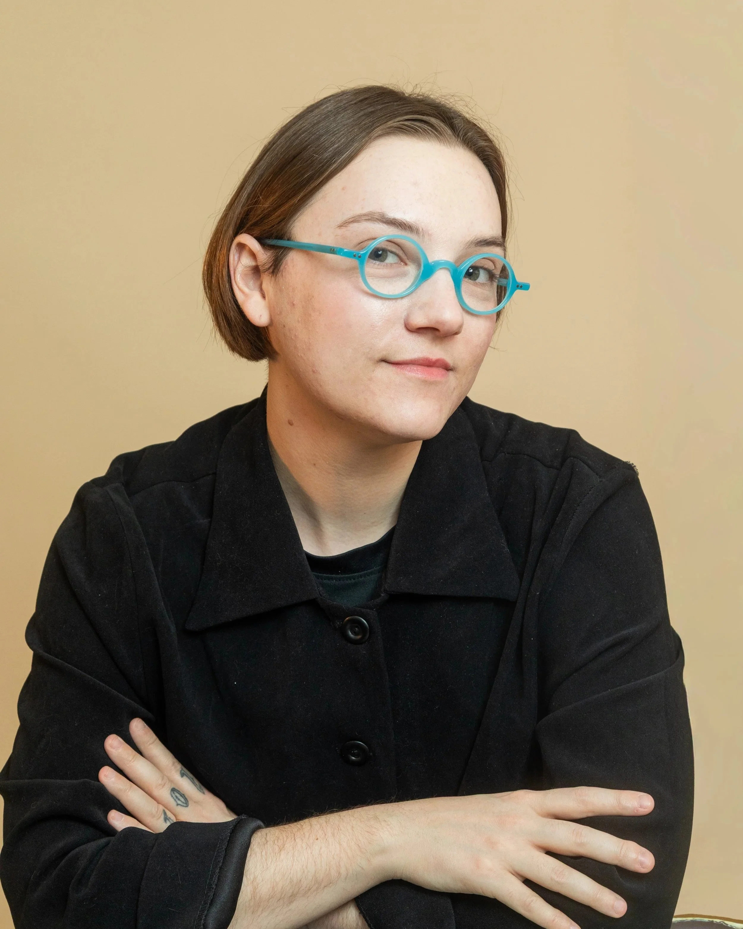 A person with short brown hair, blue glasses, and tattoos on their fingers is sitting against a beige background. They are wearing a black jacket and have their left hand on their right arm, with their right arm resting on a surface.