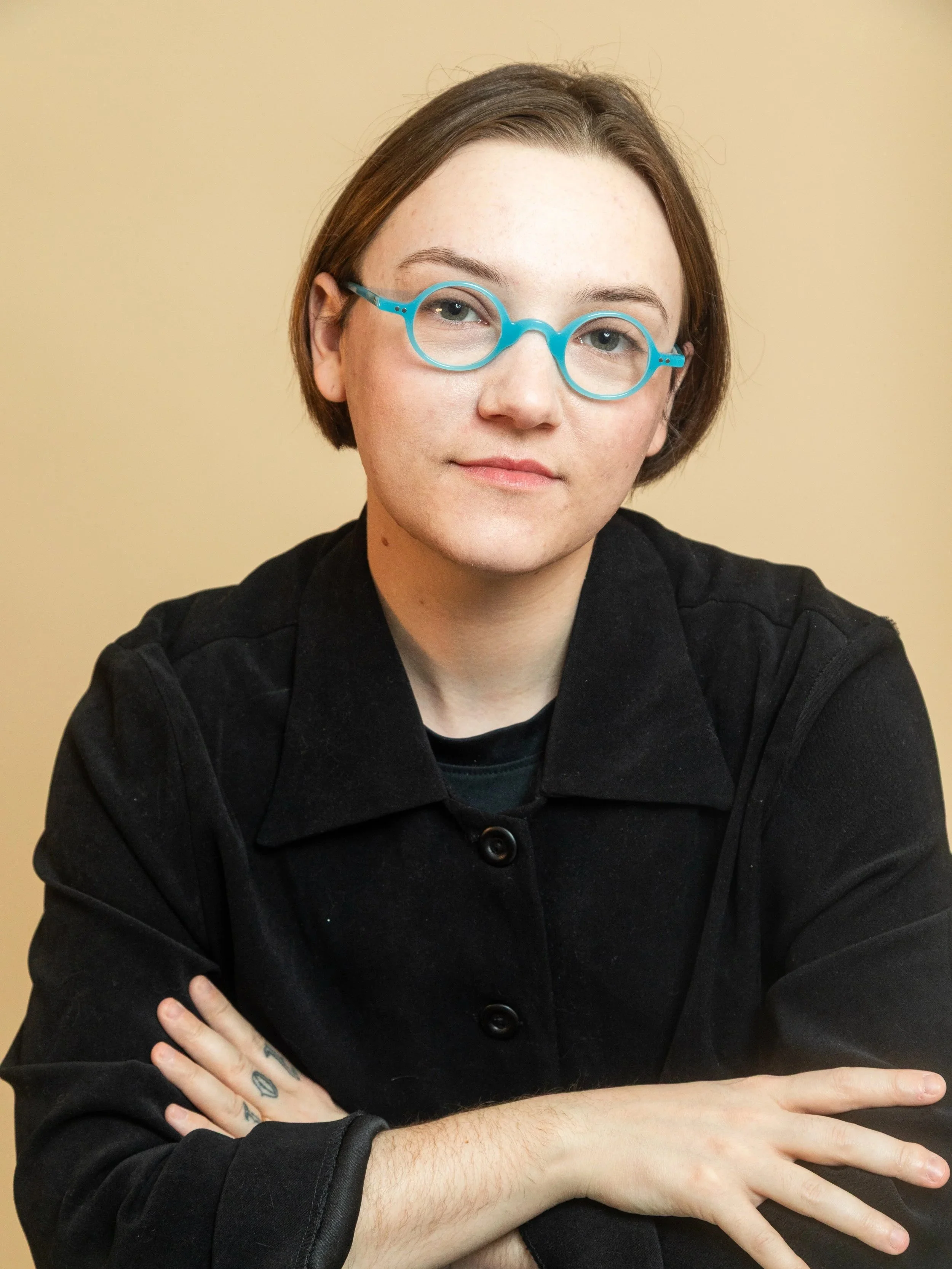 A young woman with short brown hair and teal glasses, wearing a black jacket, sitting with her arms crossed on a table against a beige background.