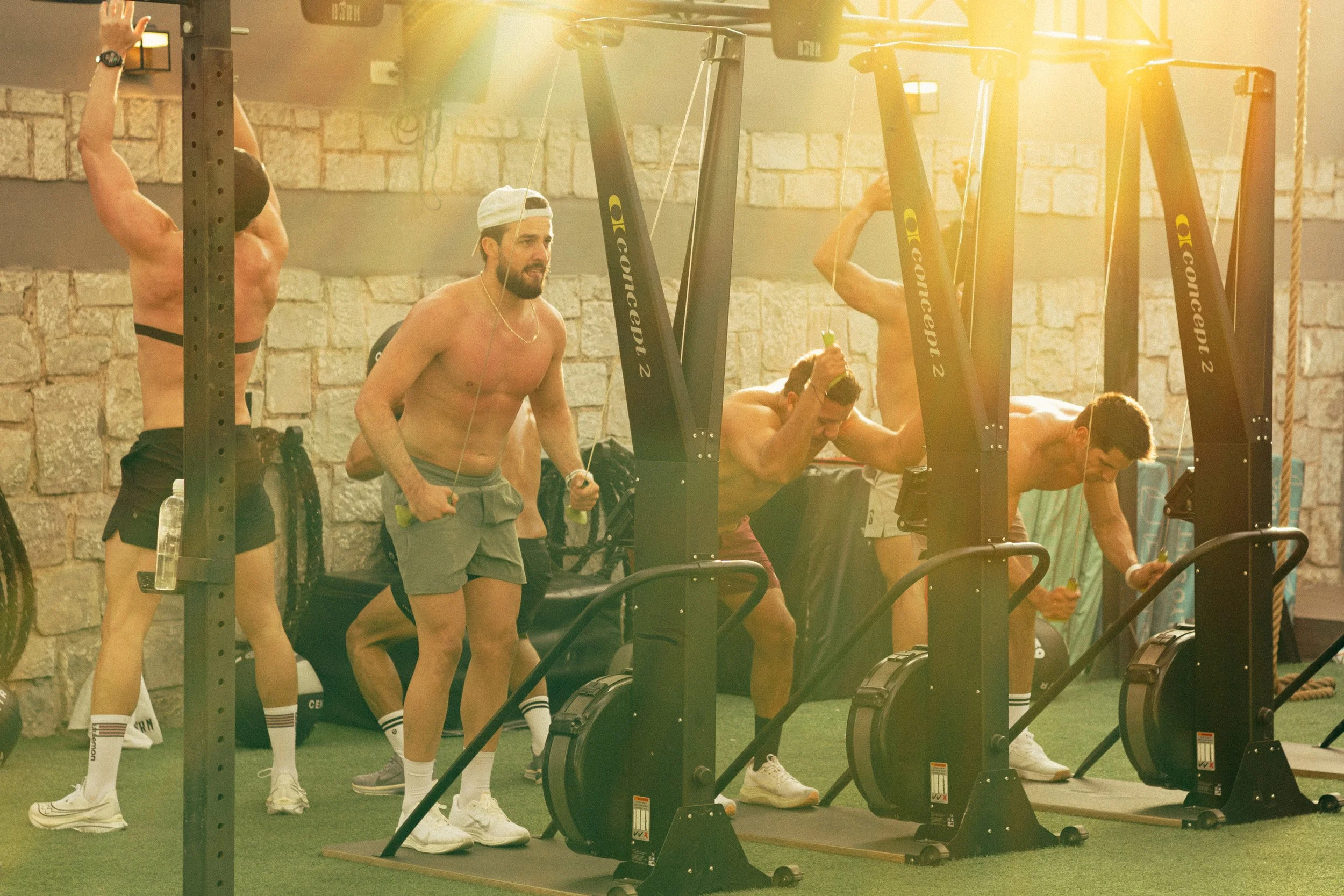 A group of men exercising at a gym, some on rowing machines and others preparing to use weights, in a brightly lit space with stone walls.