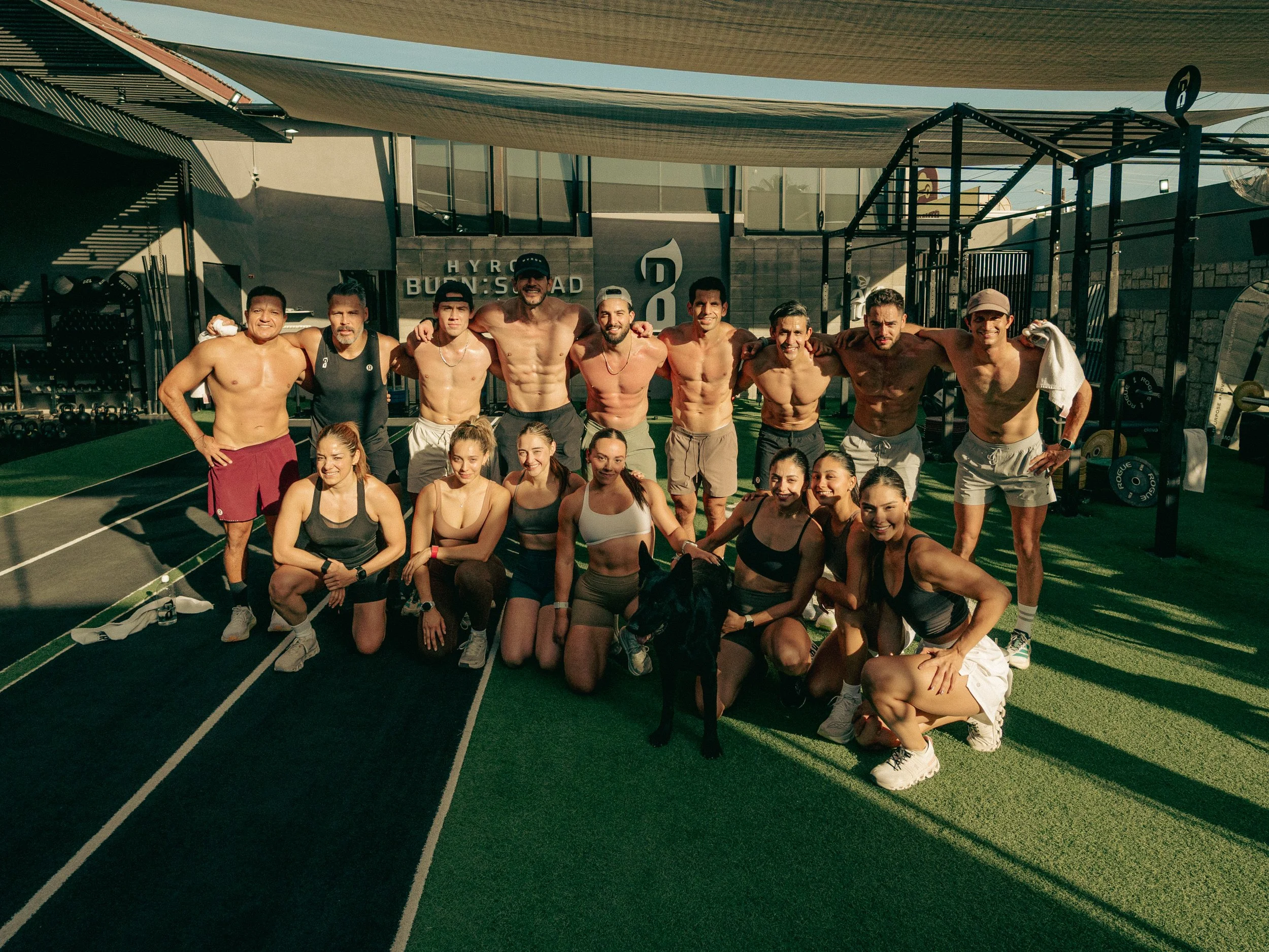 Group of ten fit, shirtless men and women smiling and posing together on a track in a gym, with workout equipment and a shaded area behind them.