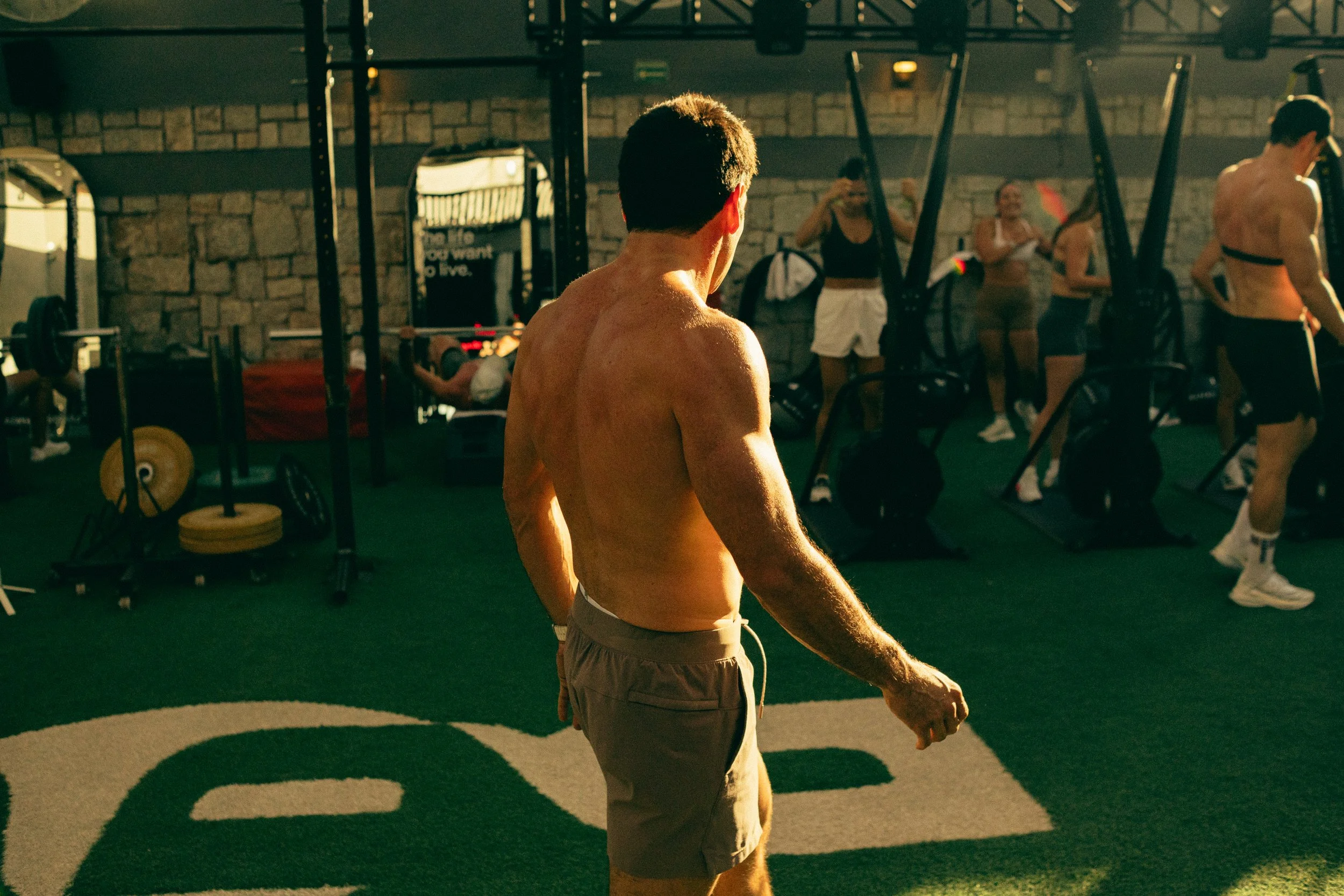 A shirtless man in a gym or workout space, with other people exercising in the background, including women and men, near gym equipment like weight racks and bars, with brick walls and motivational posters.