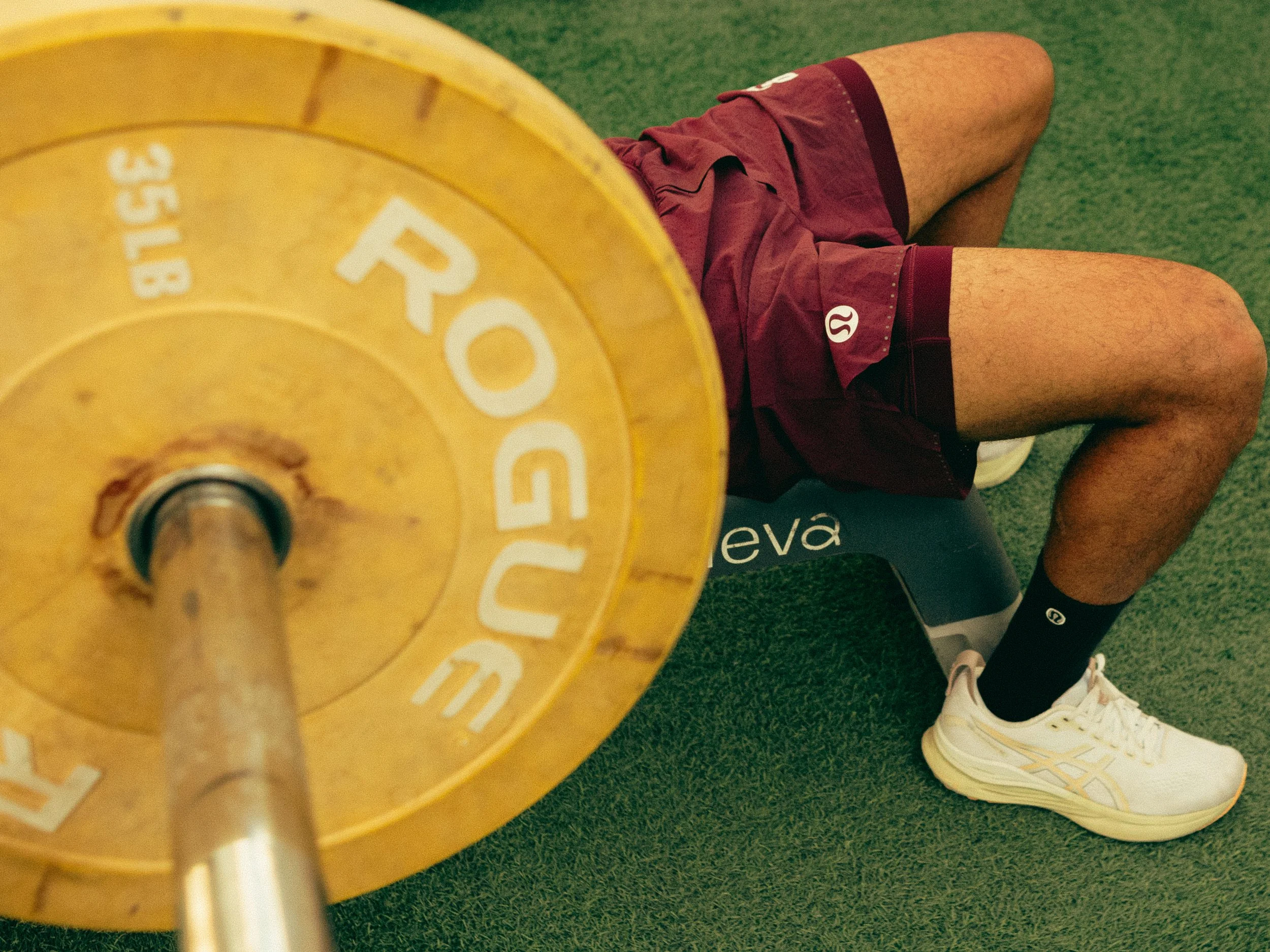 A person lying on their side on green gym flooring, wearing maroon athletic shorts and white sneakers, with a yellow weight plate held in the foreground.