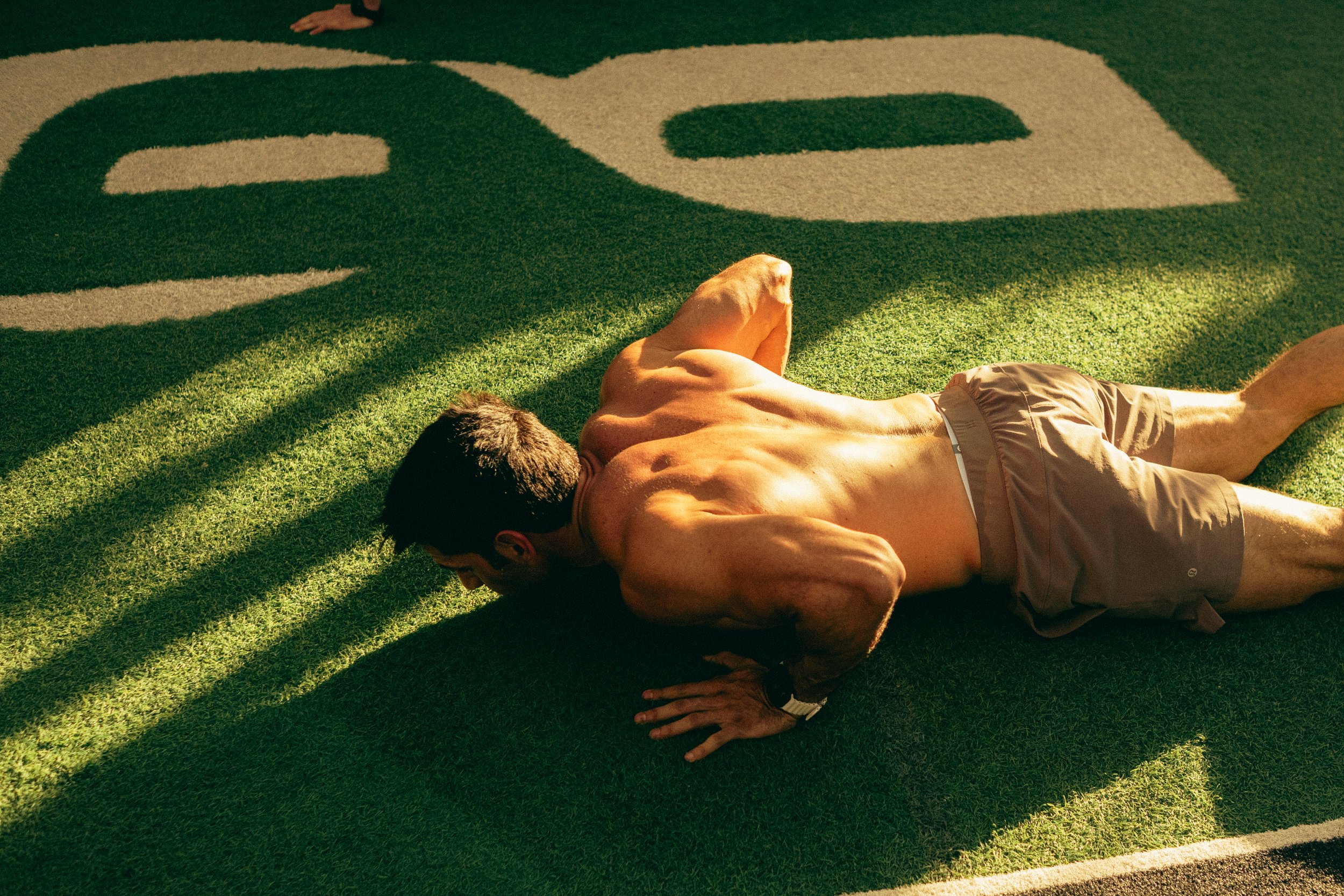 A shirtless man lying on his side on a green and beige patterned outdoor rug, with his arm raised and head resting on his arm.