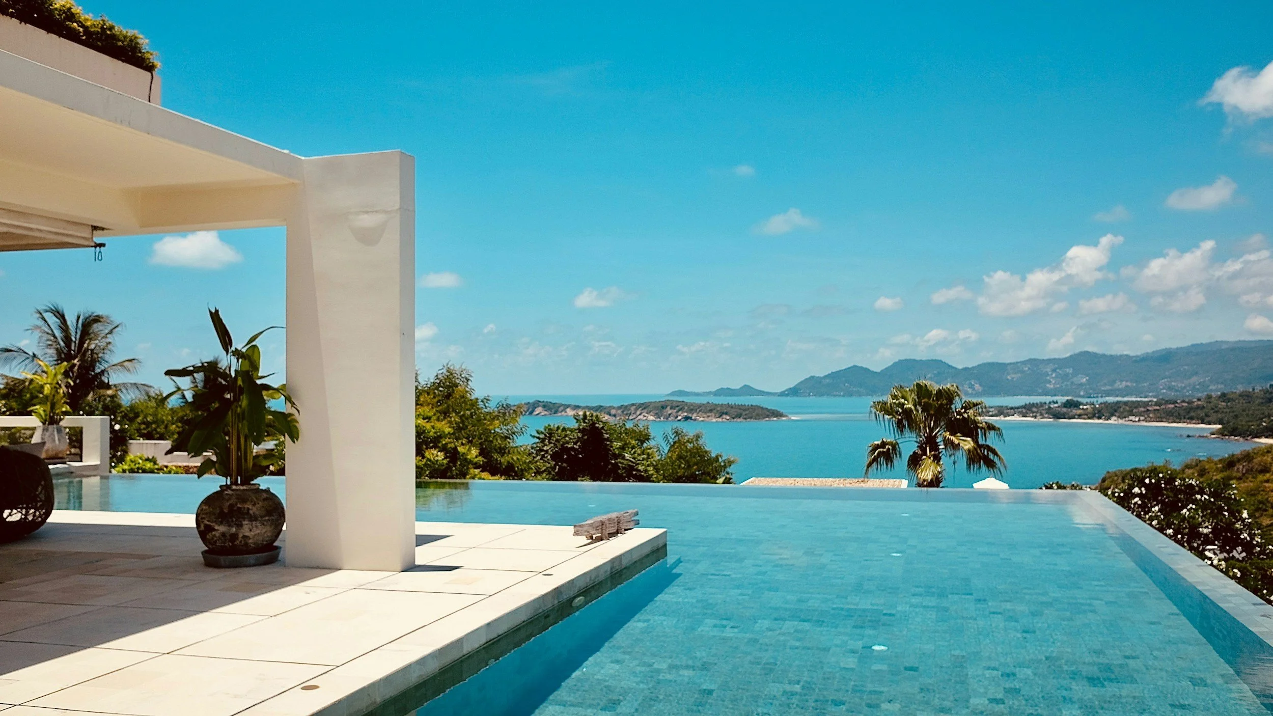 Luxury infinity pool with a view of a bay, mountains, and blue sky, with modern white architecture and green plants.