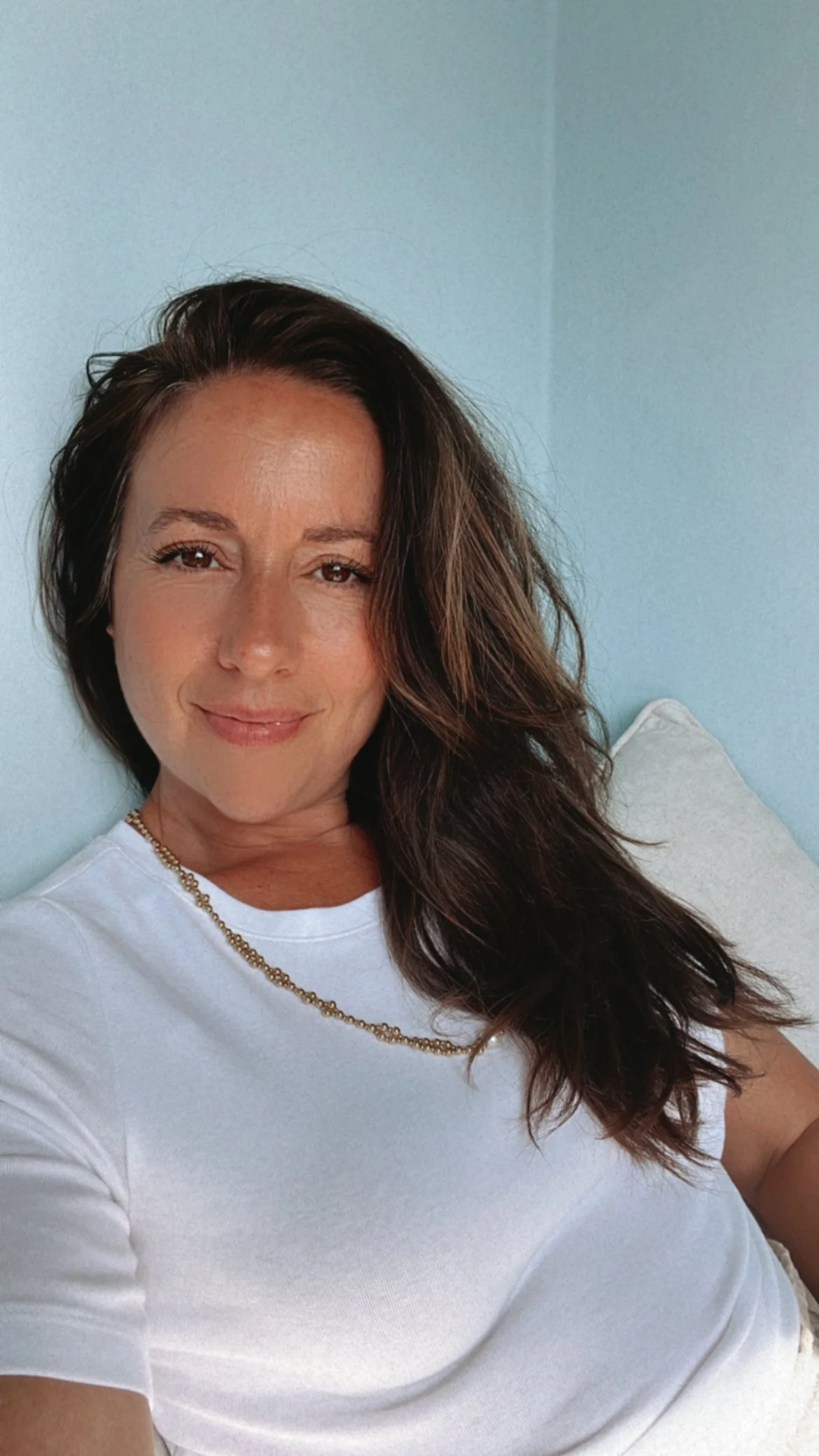 A woman with long, wavy brown hair, smiling, wearing a white t-shirt and gold chain necklace, sitting against a light blue wall.