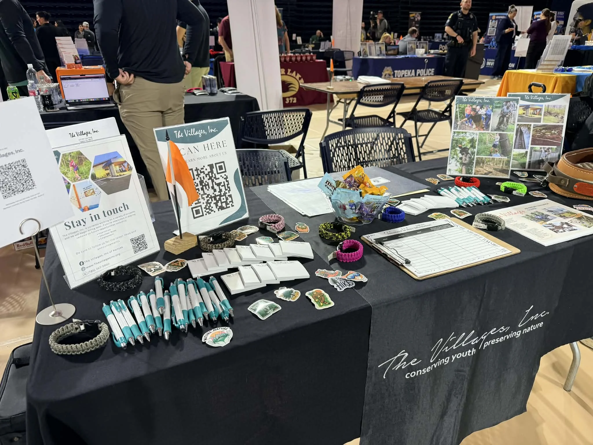 Table at an event with promotional materials including stickers, pens, and bookmarks for The Villages Inc., a youth conservation organization. There are QR codes, informational signs, and an array of small items on the table, with chairs around and b