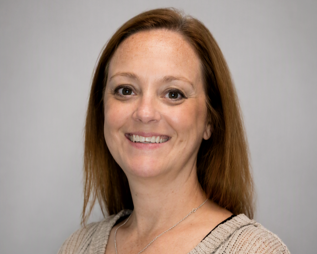 Smiling woman with shoulder-length red hair, fair skin, and freckles, wearing a beige knit sweater and a necklace, standing against a plain gray background.