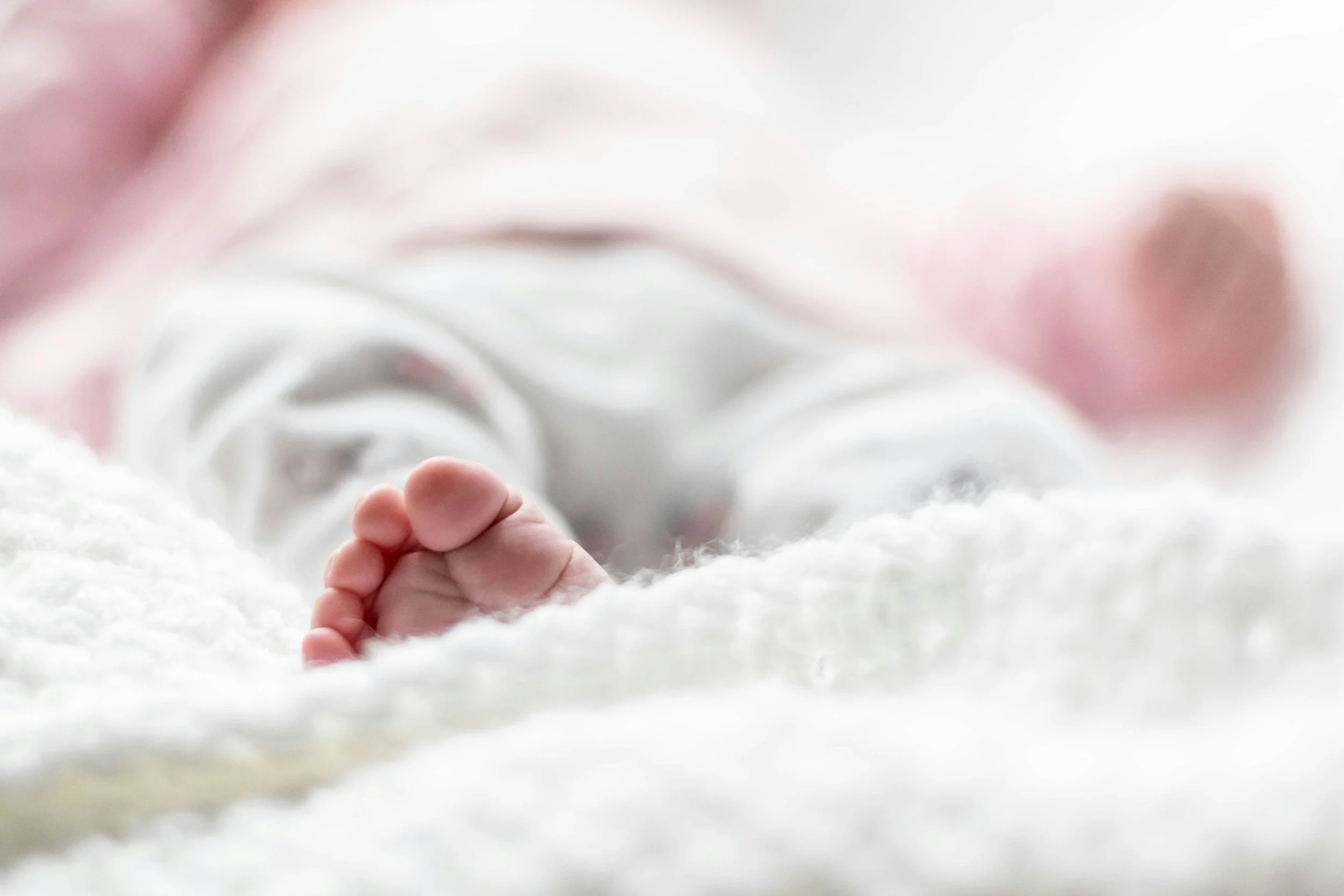 Close-up of a baby's tiny hand gripping a soft white blanket with pink accents, lying on a plush white surface.