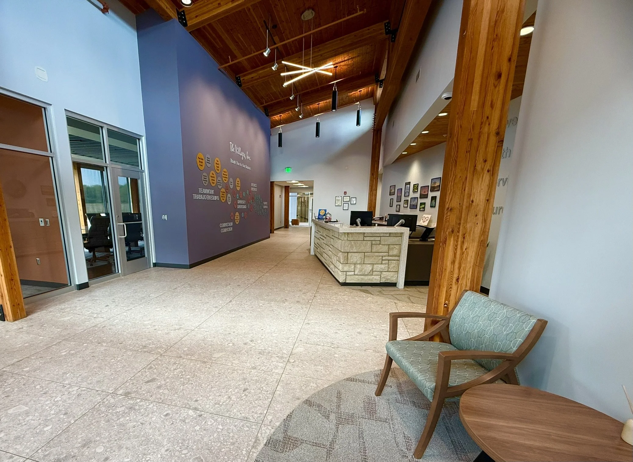 The interior of a modern office lobby with wooden beams and ceiling, a reception desk made of stone, and a seating area with two wooden chairs with patterned cushions. The wall behind the desk features framed pictures and a purple accent wall with writing and colorful circles.