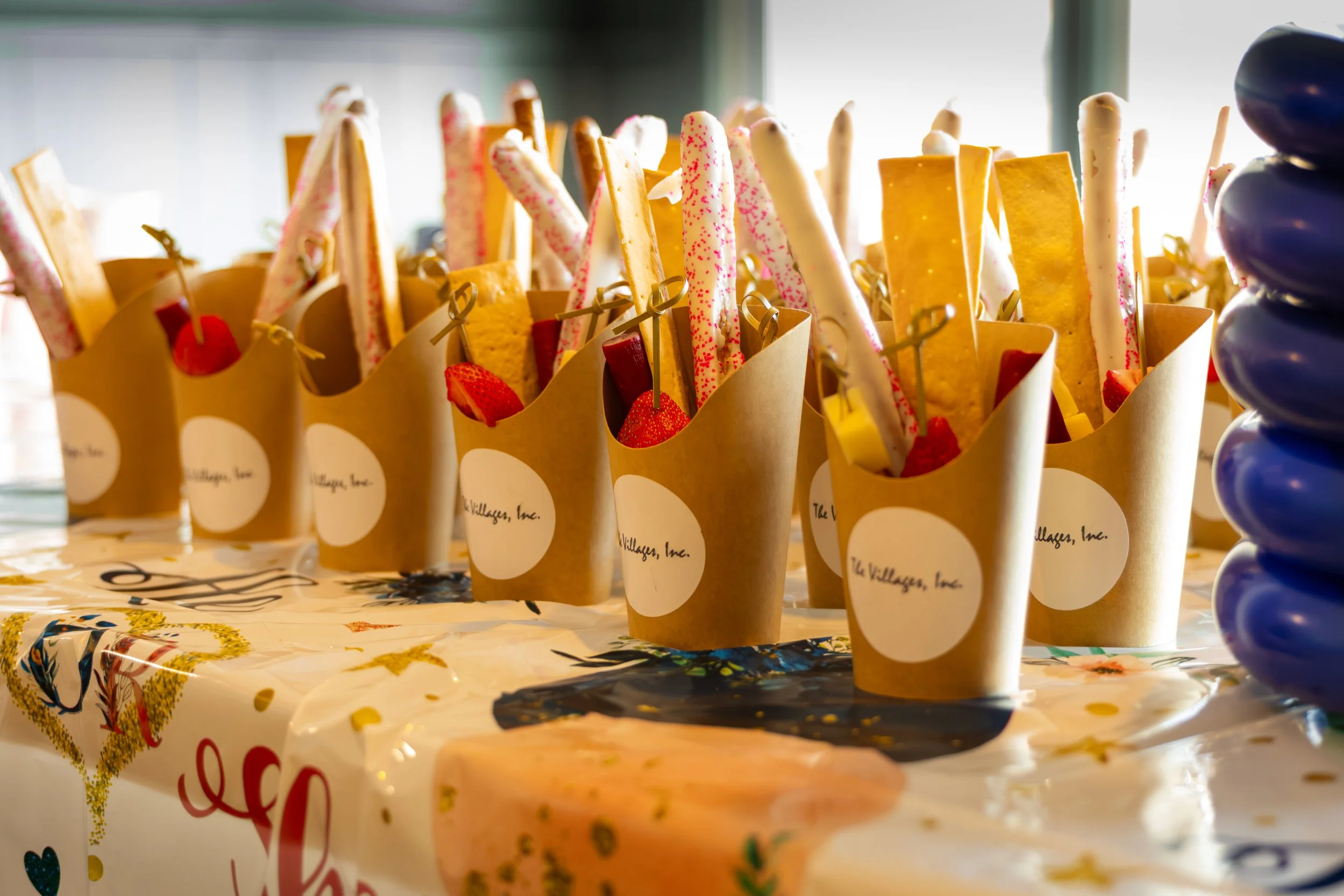 Multiple paper cones filled with decorated cookies, strawberries, and candy sticks on a table with a decorative tablecloth.