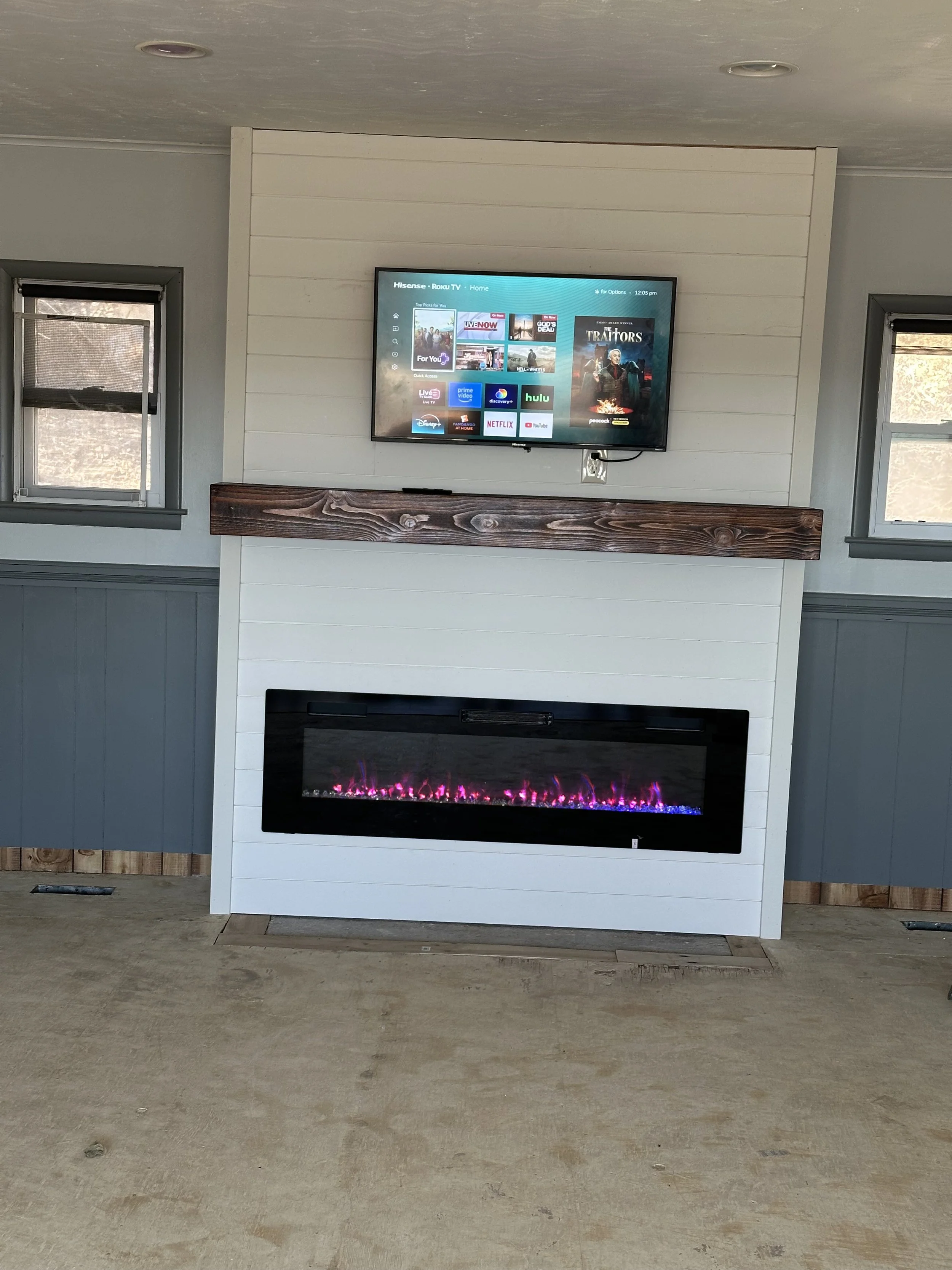 A living room with two small windows on either side of a wall-mounted television. Below the television is a fireplace with a white mantel and a colorful electric fire display. The room has a unfinished wooden floor and walls painted in shades of gray