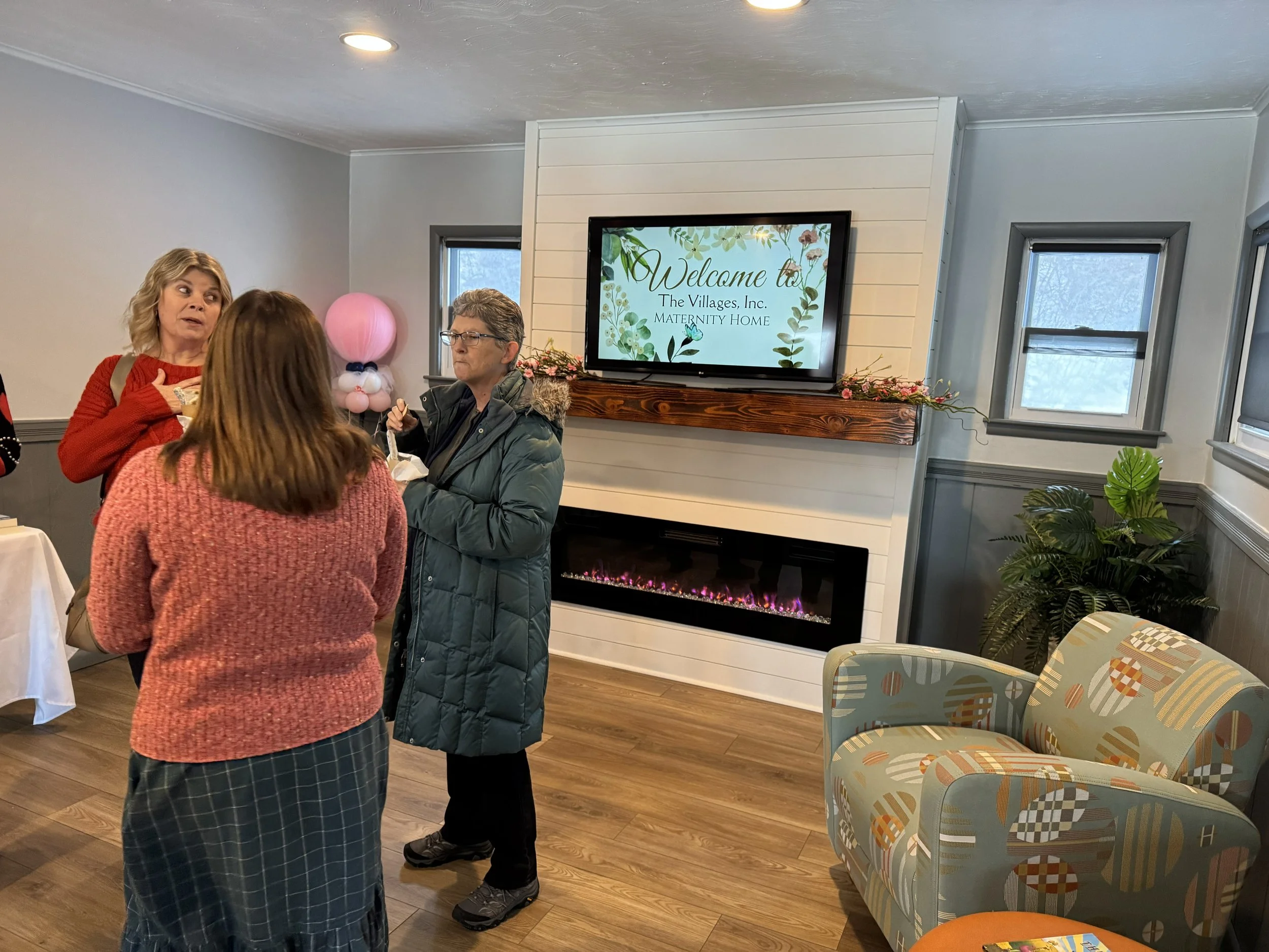 Three women are talking in a living room near a wall with a large flat-screen TV displaying a welcome message for The Villages Inc. Maternity Home. The room has a white wall and a fireplace, plants, and windows.