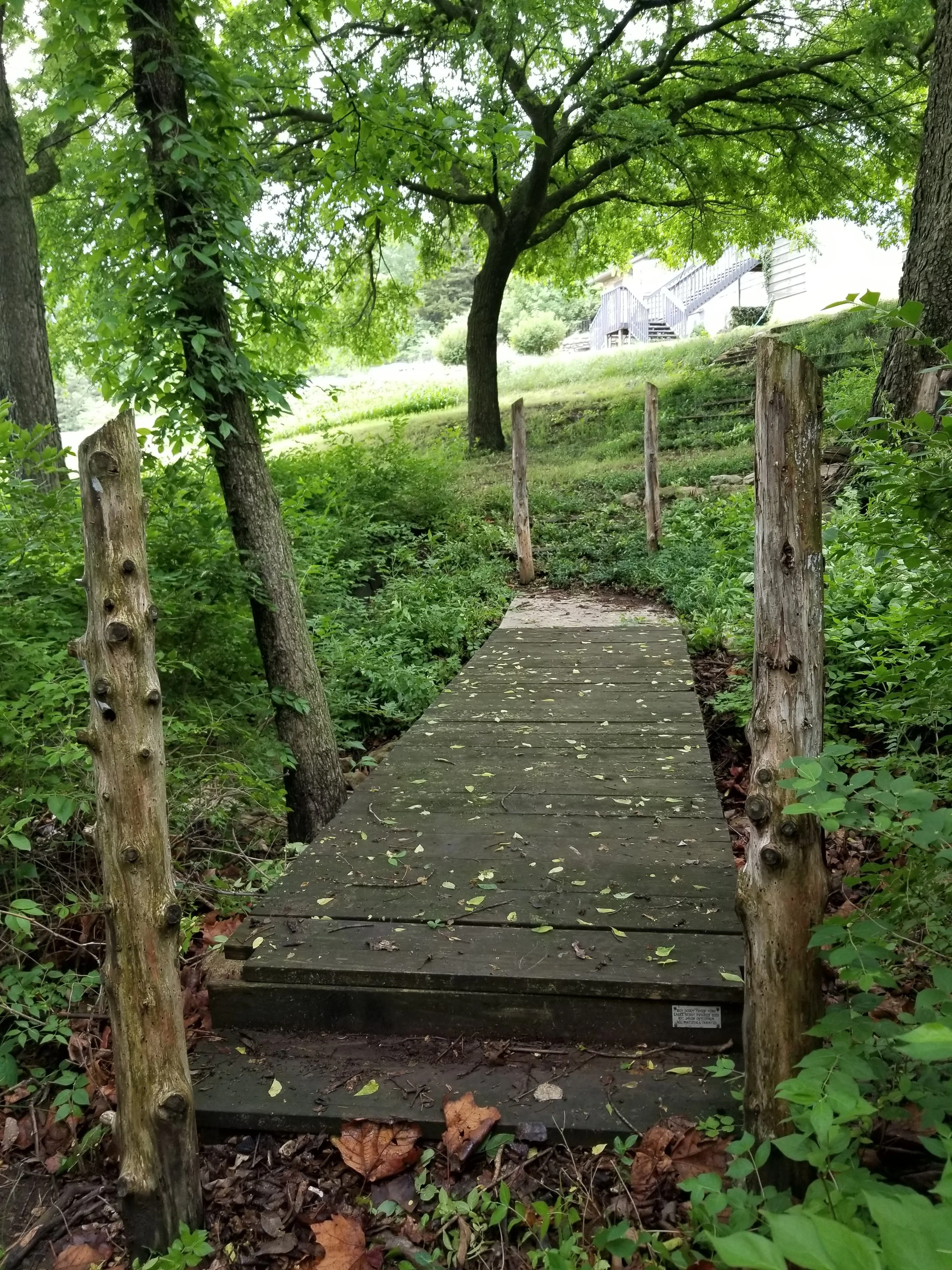 A narrow wooden footbridge crossing over a small stream in a lush, green backyard garden with trees and bushes. A house with stairs and a deck is visible in the background.