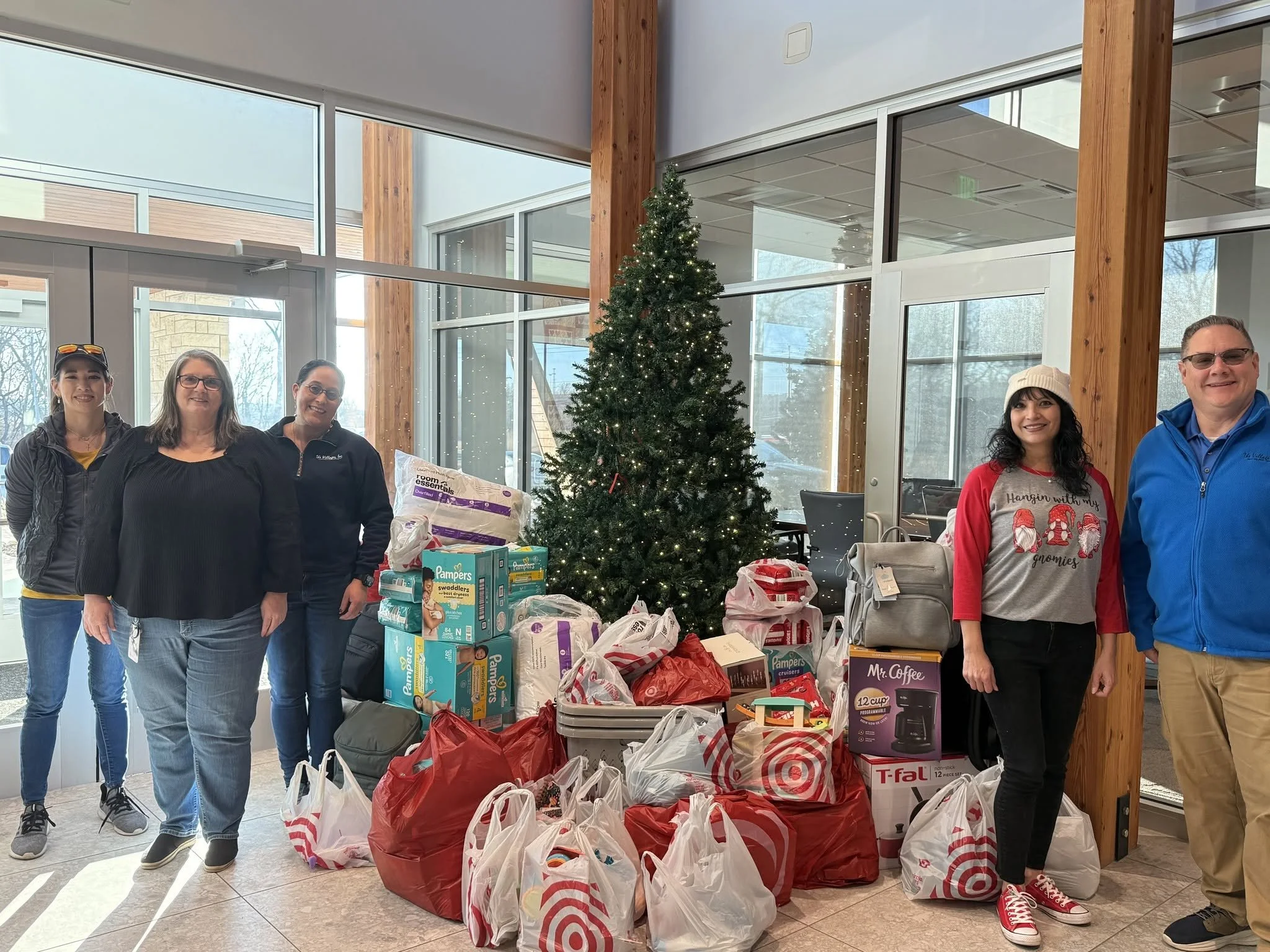 A group of five people standing inside a building with a large Christmas tree decorated with lights. They are surrounded by bags of donations and boxes of supplies. The people are smiling at the camera.