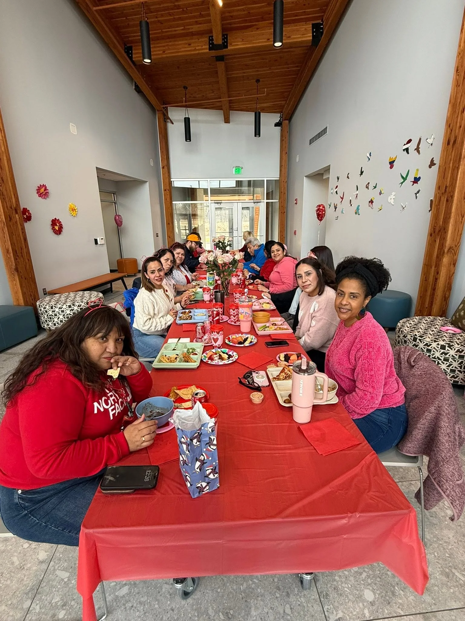 A group of women sitting around a long table with red tablecloth, enjoying a meal together at a celebration or gathering.