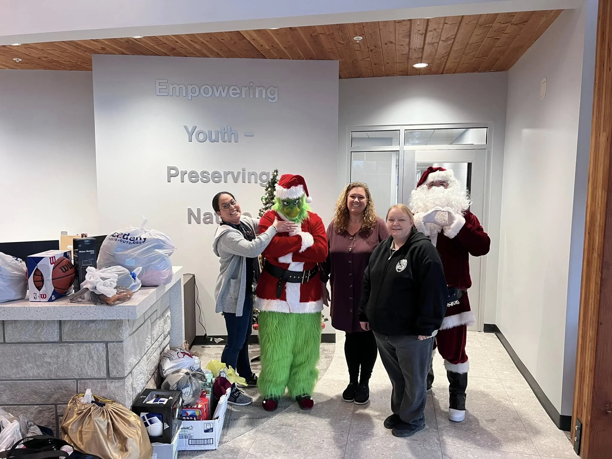 People posing with a person dressed as the Grinch and Santa Claus in a room with a Christmas tree and donations.