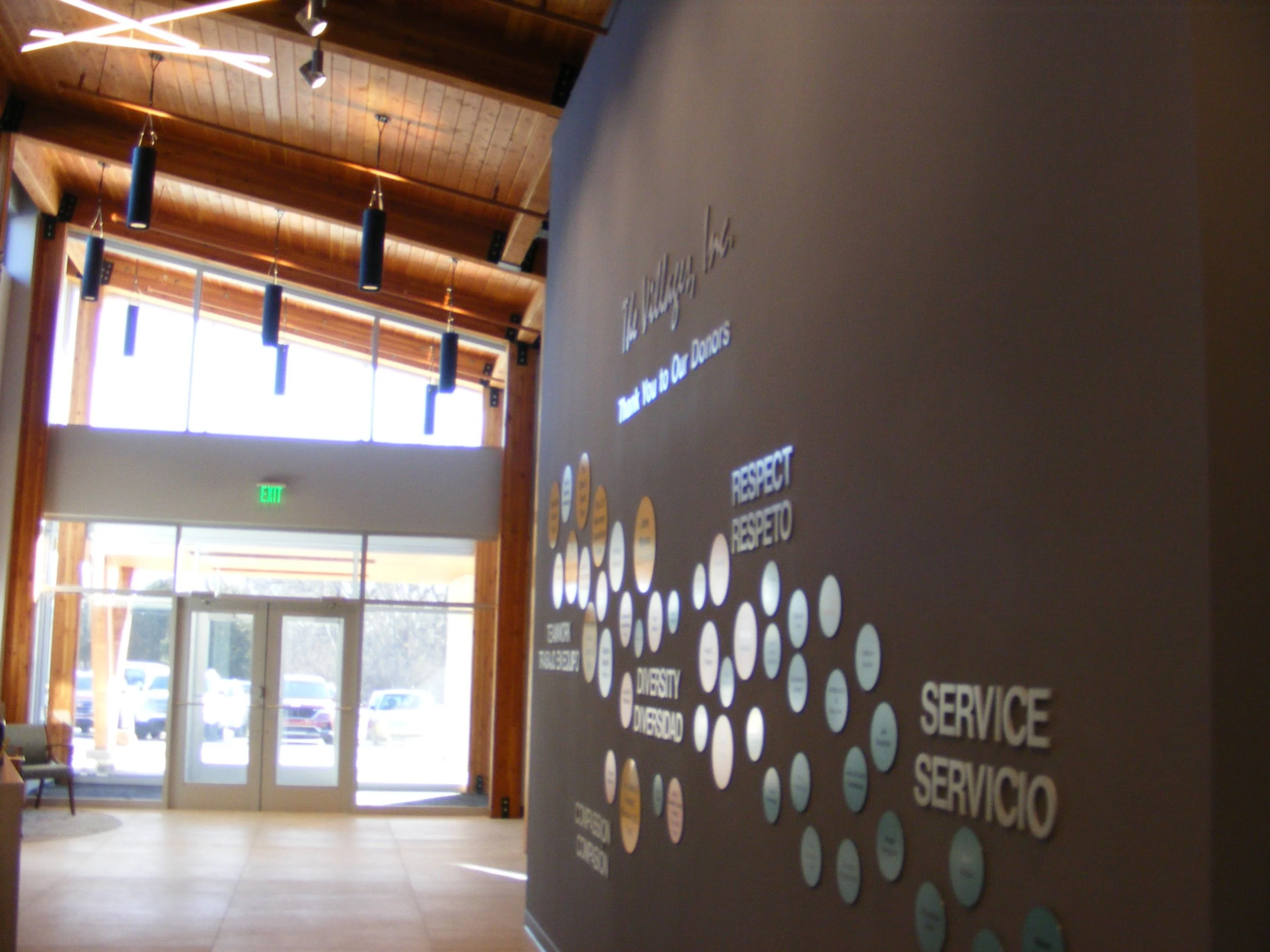 Interior of a building with wooden accent ceiling, hanging black pendant lights, glass entrance doors, and a wall with circular motivational words including respect, dignity, service, etc.