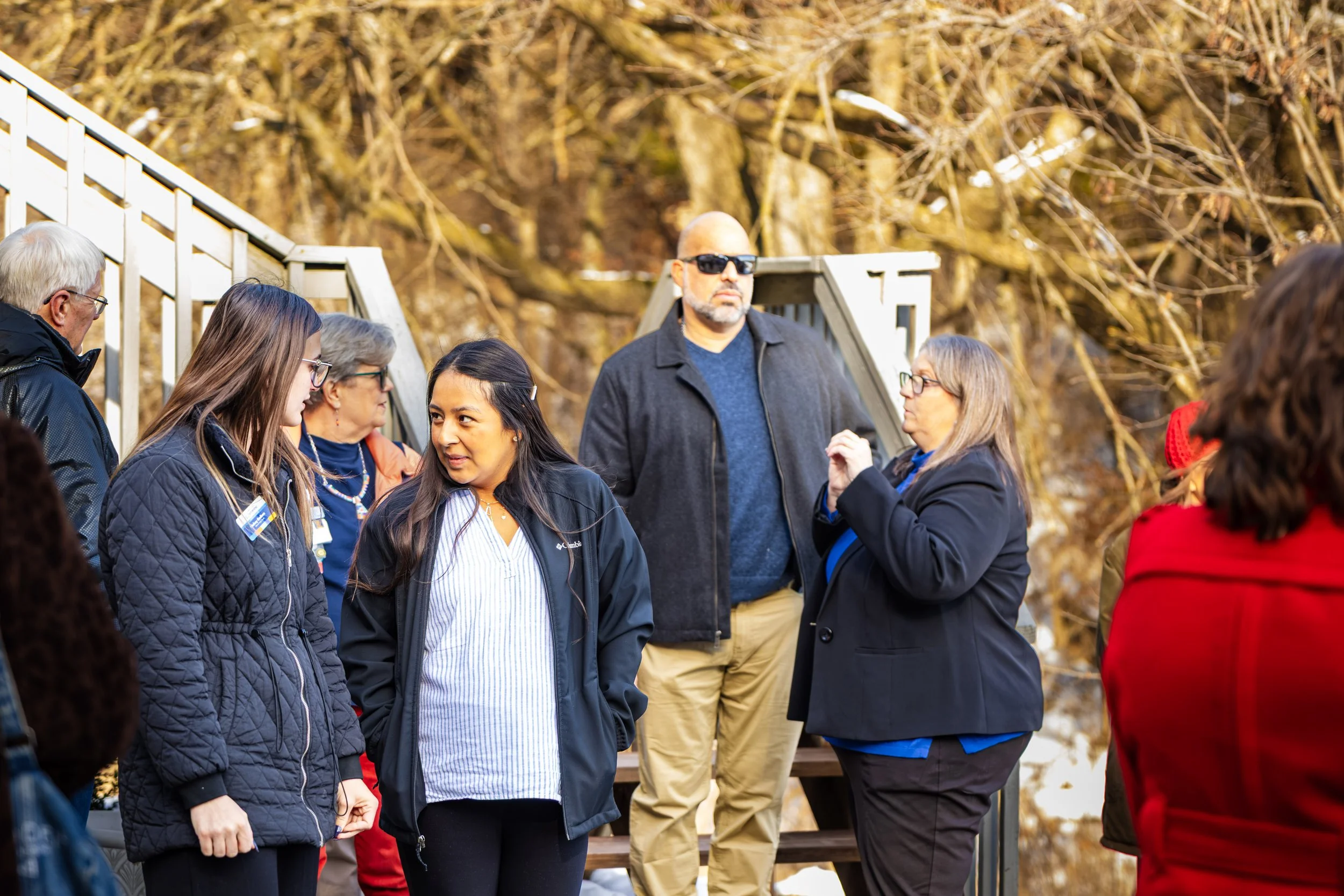 A group of people, including women and men, are gathered outdoors on a sunny day, engaging in conversation with a background of leafless trees.