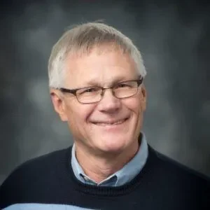 Headshot of a smiling man with gray hair wearing glasses, a blue collared shirt, and a dark sweater against a blurred background.