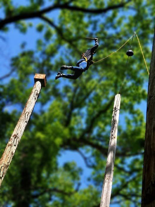 A person in a harness and helmet is jumping off a platform between two tall wooden poles, with safety ropes, in an outdoor adventure course surrounded by green trees.