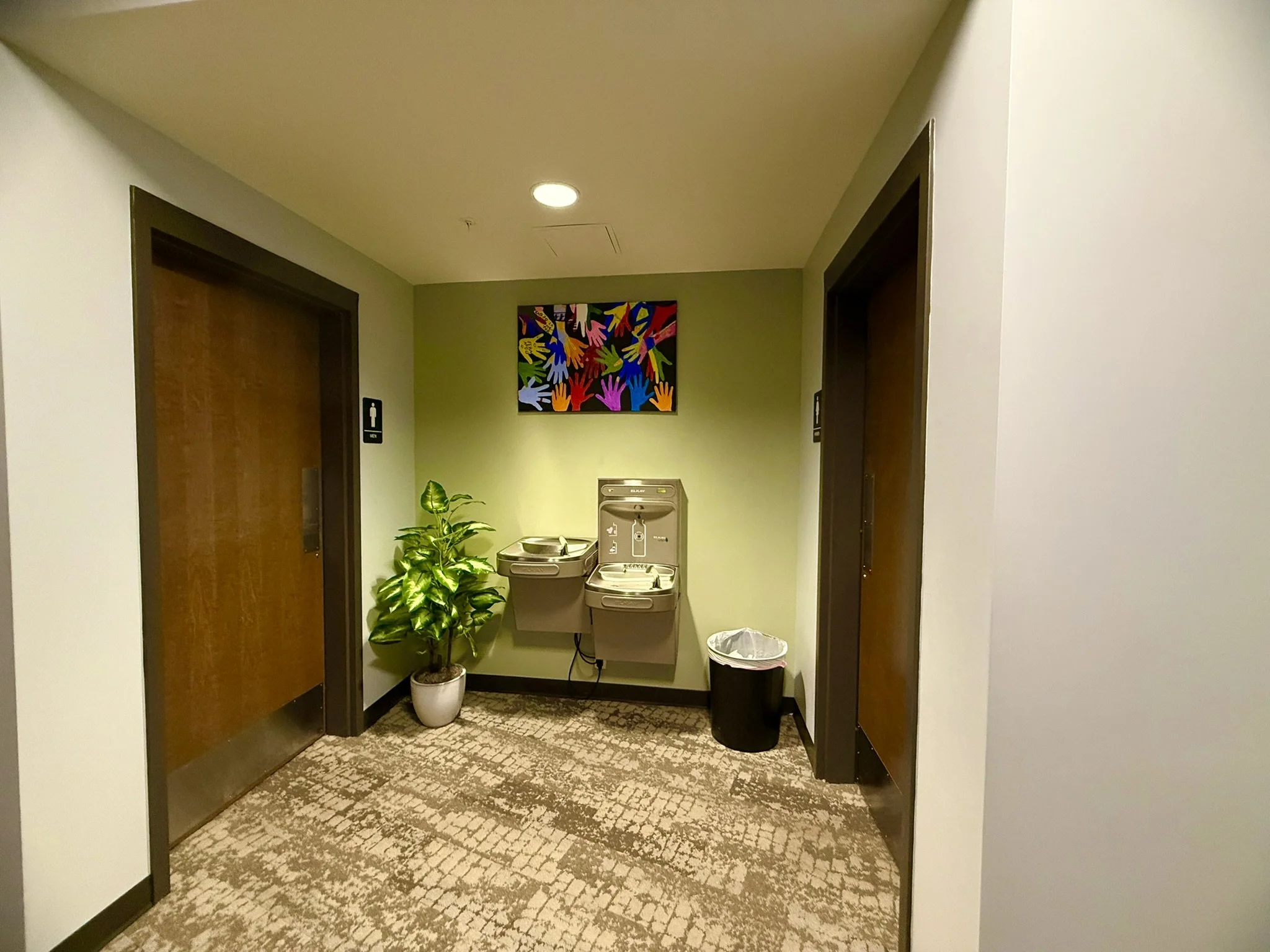 A hallway with two wooden doors, one of which has a men's restroom sign. There is a colorful abstract artwork on the back wall, along with a water fountain, a potted plant, a trash can, and a carpeted floor.