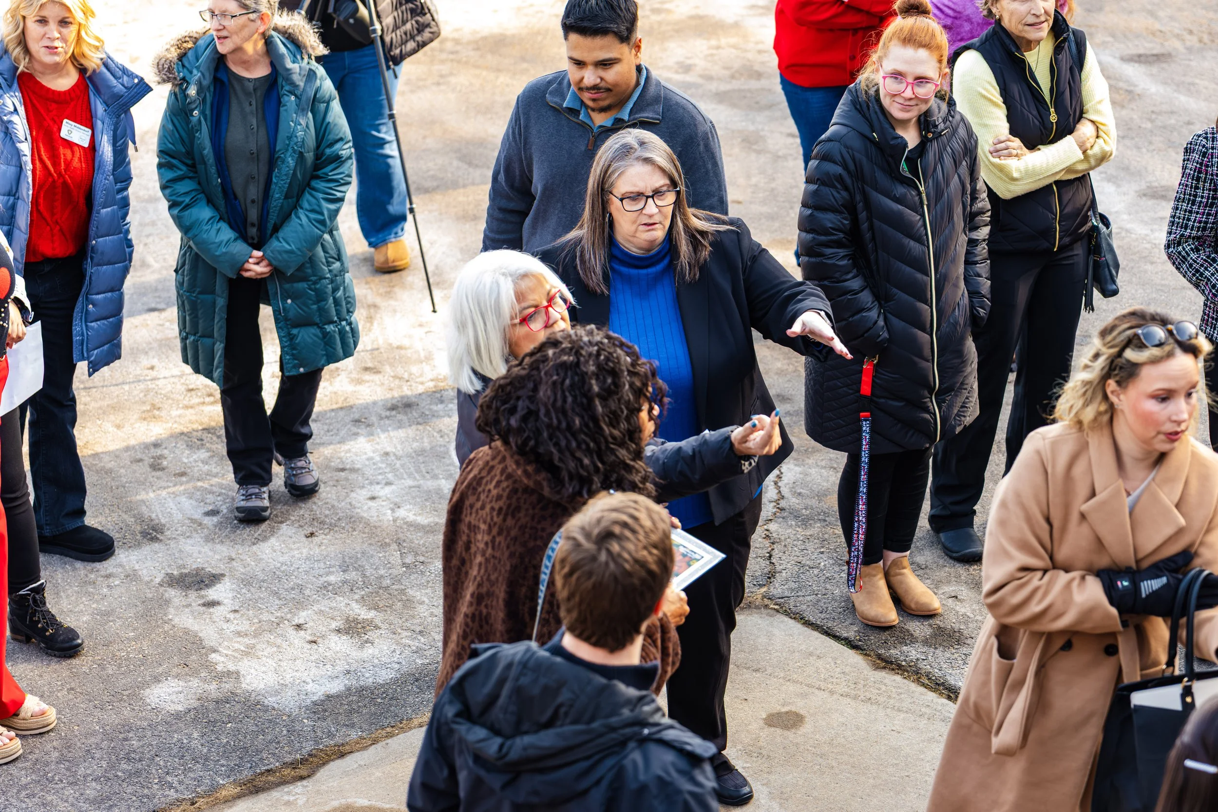 A diverse group of people standing outdoors, with some seated and others standing, engaged in a discussion or tour, dressed warmly for cool weather.