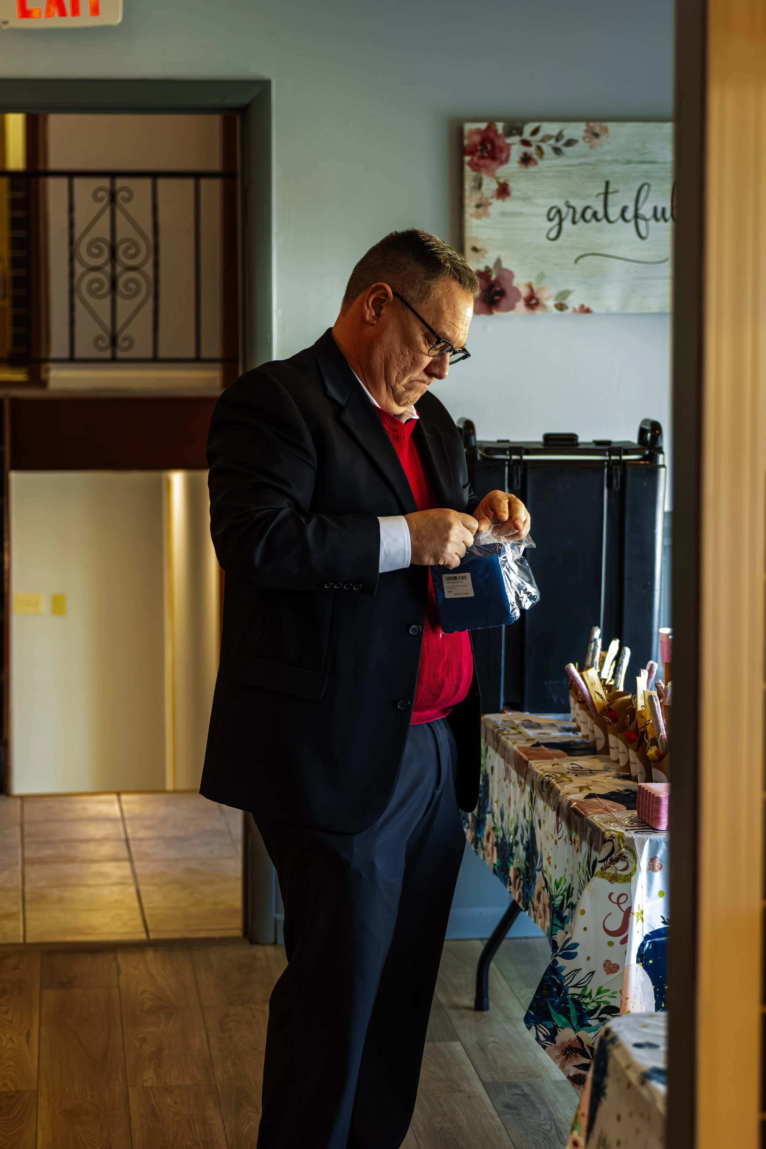 A man with glasses wearing a black suit jacket and a red sweater looking at items on a table with a colorful tablecloth, inside a room with wooden floor and a framed floral and grateful message on the wall.