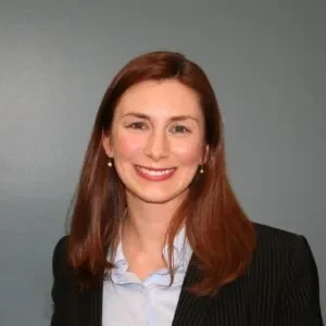 A woman with shoulder-length brown hair, wearing a black blazer and light blue shirt, smiling against a plain gray background.