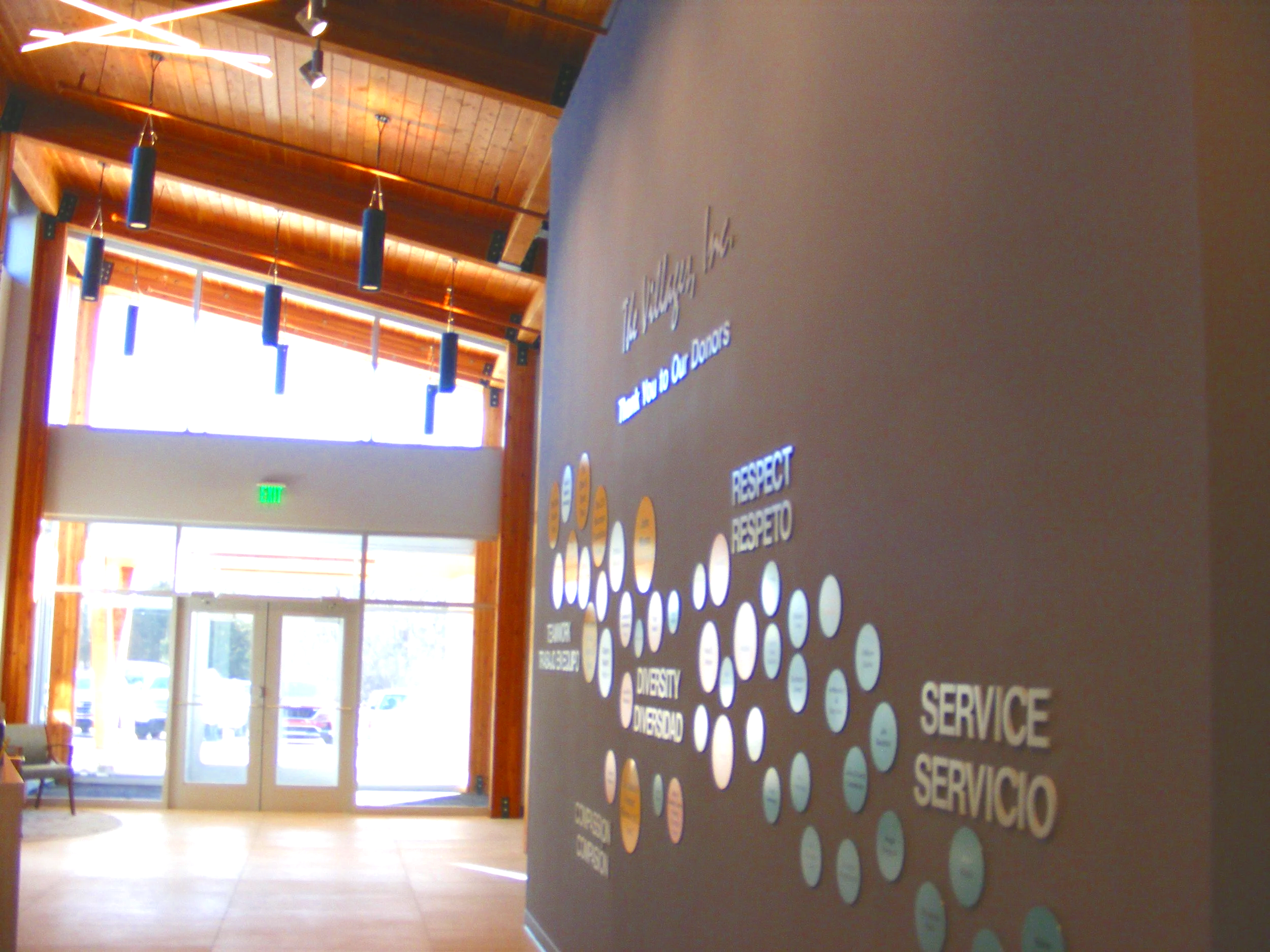 The image shows the lobby of a building with a wooden ceiling, hanging pendant lights, and large windows. There is a wall with circular labels indicating different services such as 'DIVERSIDAD,' 'RESPECT,' and 'SERVICE.'
