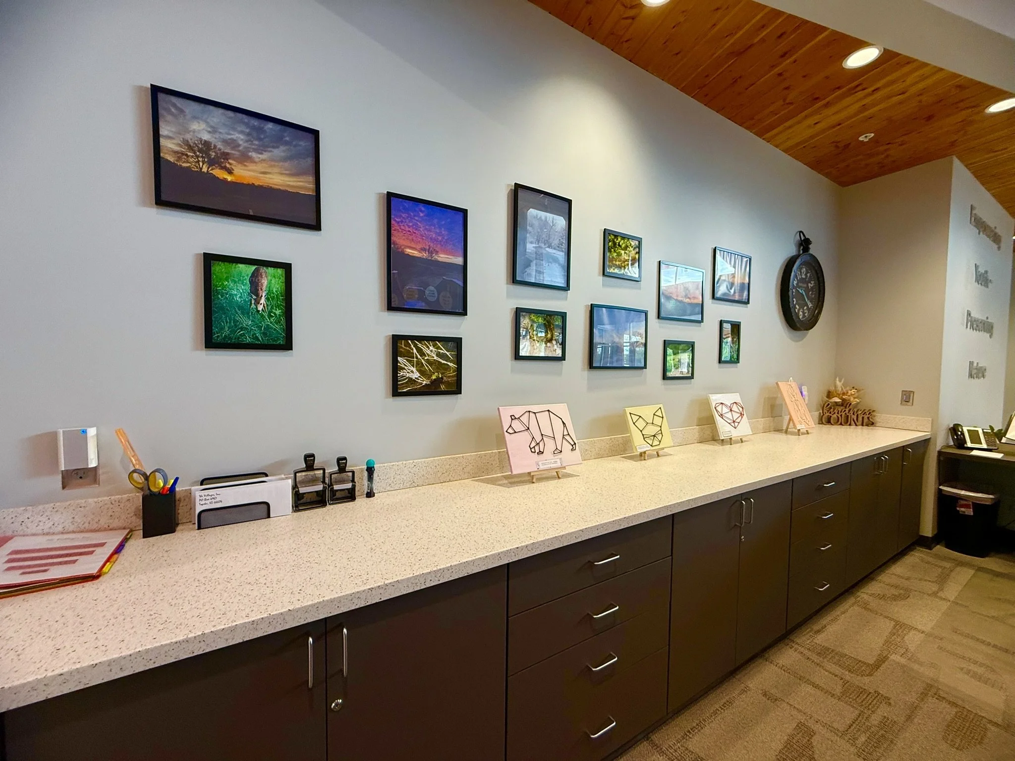 View of a reception area with a white countertop, art prints of animals and landscapes on the wall, a large clock, and various office supplies and decorations on the countertop.