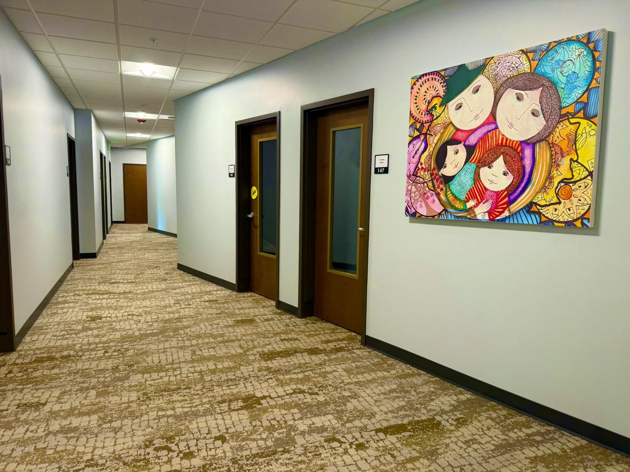 Long hallway in an office building with white walls, brown doors, and a patterned beige carpet. There is colorful abstract artwork on the wall to the right.