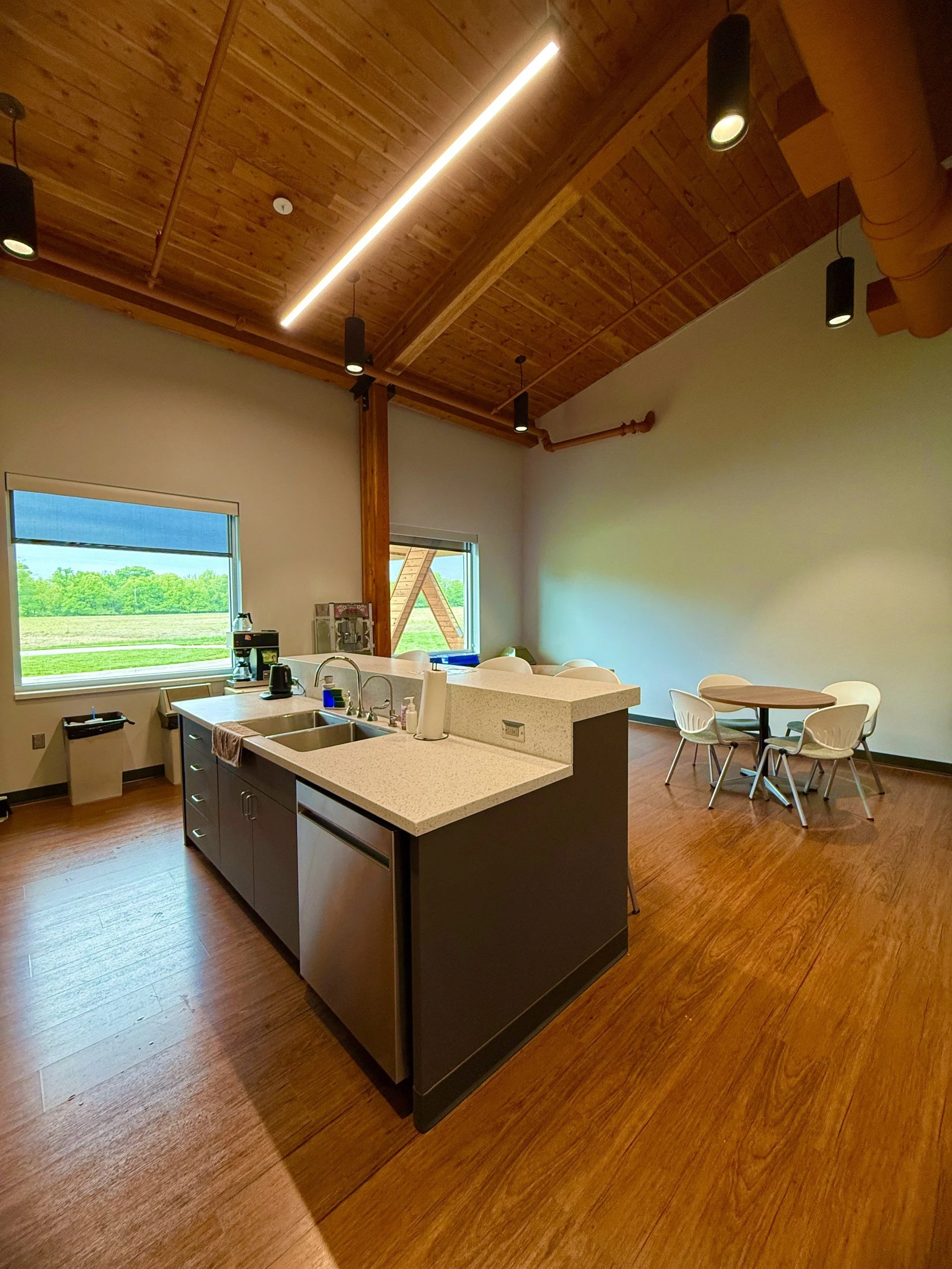 Interior view of a room with a kitchen island, white chairs around a round table, wooden flooring, a wooden ceiling with modern lighting, and two windows showing greenery outside.