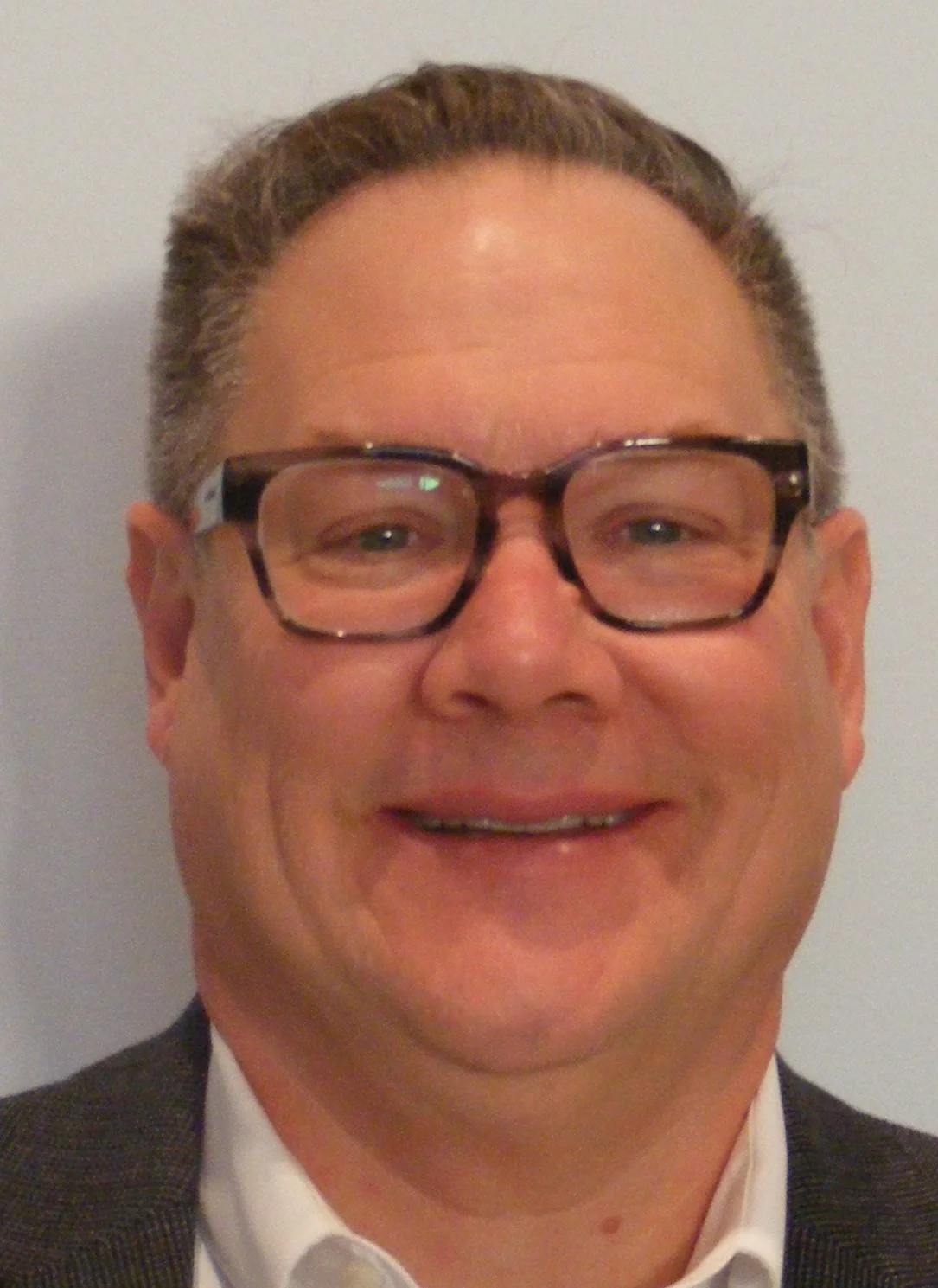 Close-up of a smiling man wearing glasses, a suit, and a tie, with light skin and short brown hair.