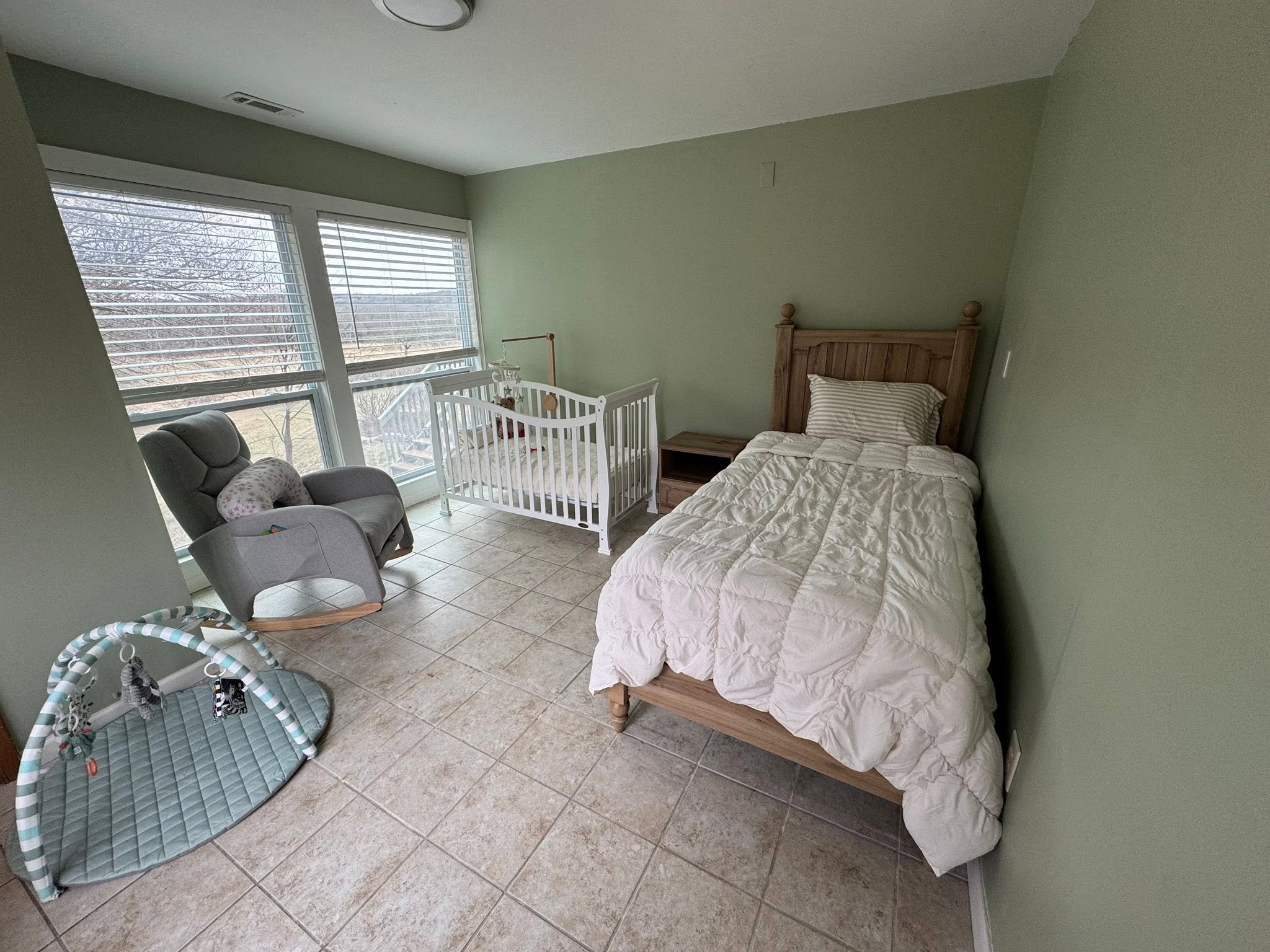 A bedroom with a twin bed, a wooden headboard, white bedding, a gray armchair, a baby crib, and a window with blinds revealing leafless trees outside.