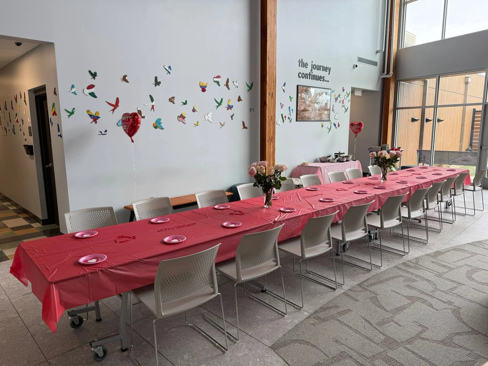 Empty banquet table with pink tablecloth, pink plates, and vases with pink roses, decorated for a celebration, with wall decor including colorful paper birds and a sign that reads "the journey continues..." in a bright, airy room with large windows.