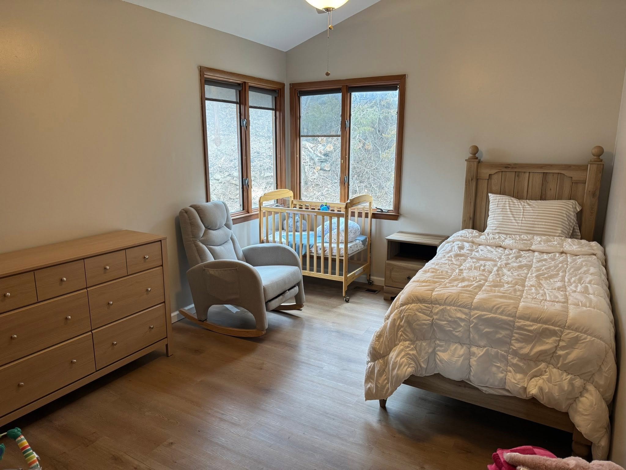 A bedroom with wooden flooring, a large bed with white textured bedding, a wooden headboard, a nightstand, a gray recliner chair, a baby crib near a window, and a dresser. The window has three sections with wooden frames, showing an outside view of t