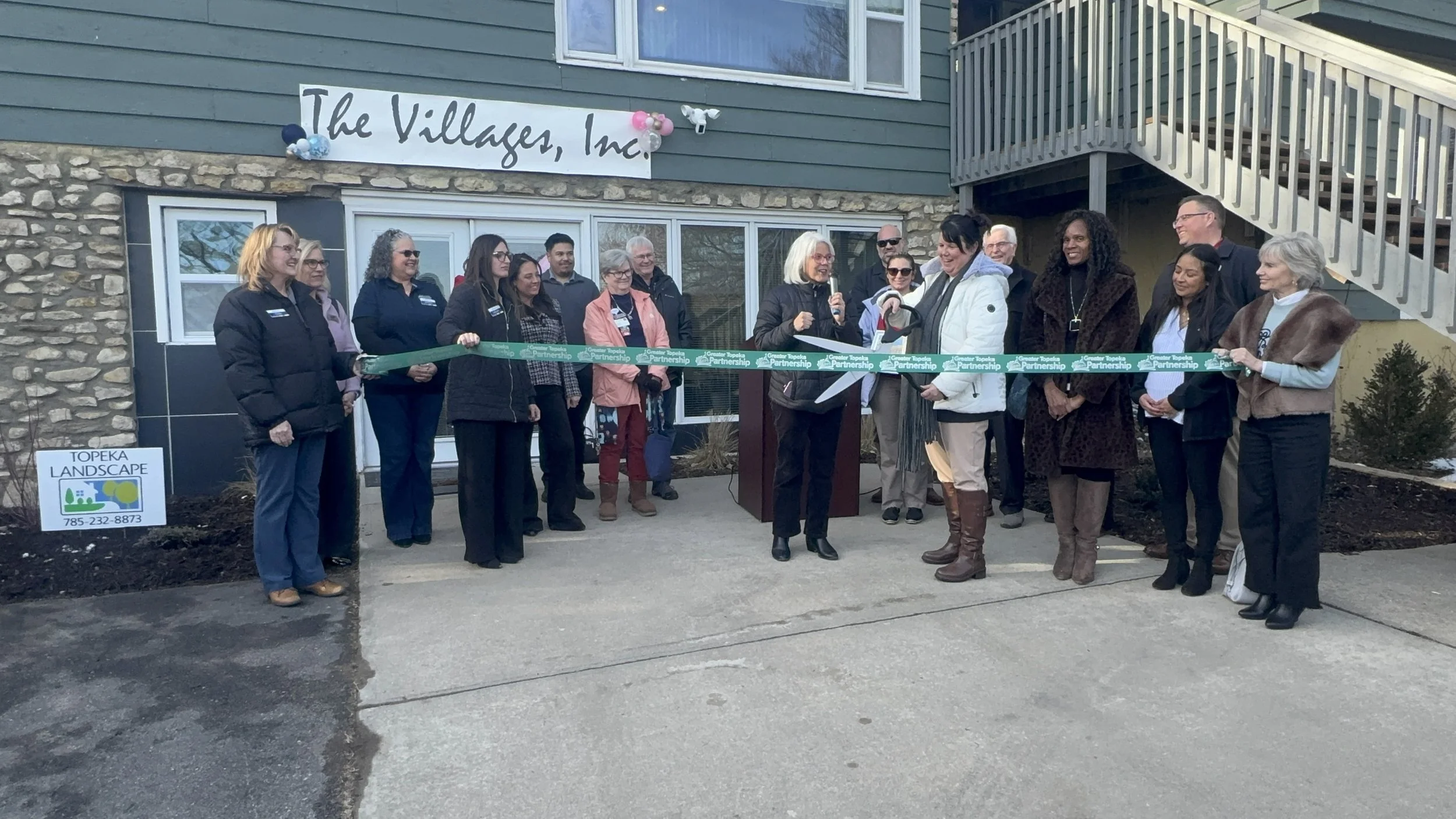 Group of people at a ribbon-cutting event outside a building with a sign that reads 'The Villages, Inc.' and a banner that says 'Greater Topeka Partnership'.