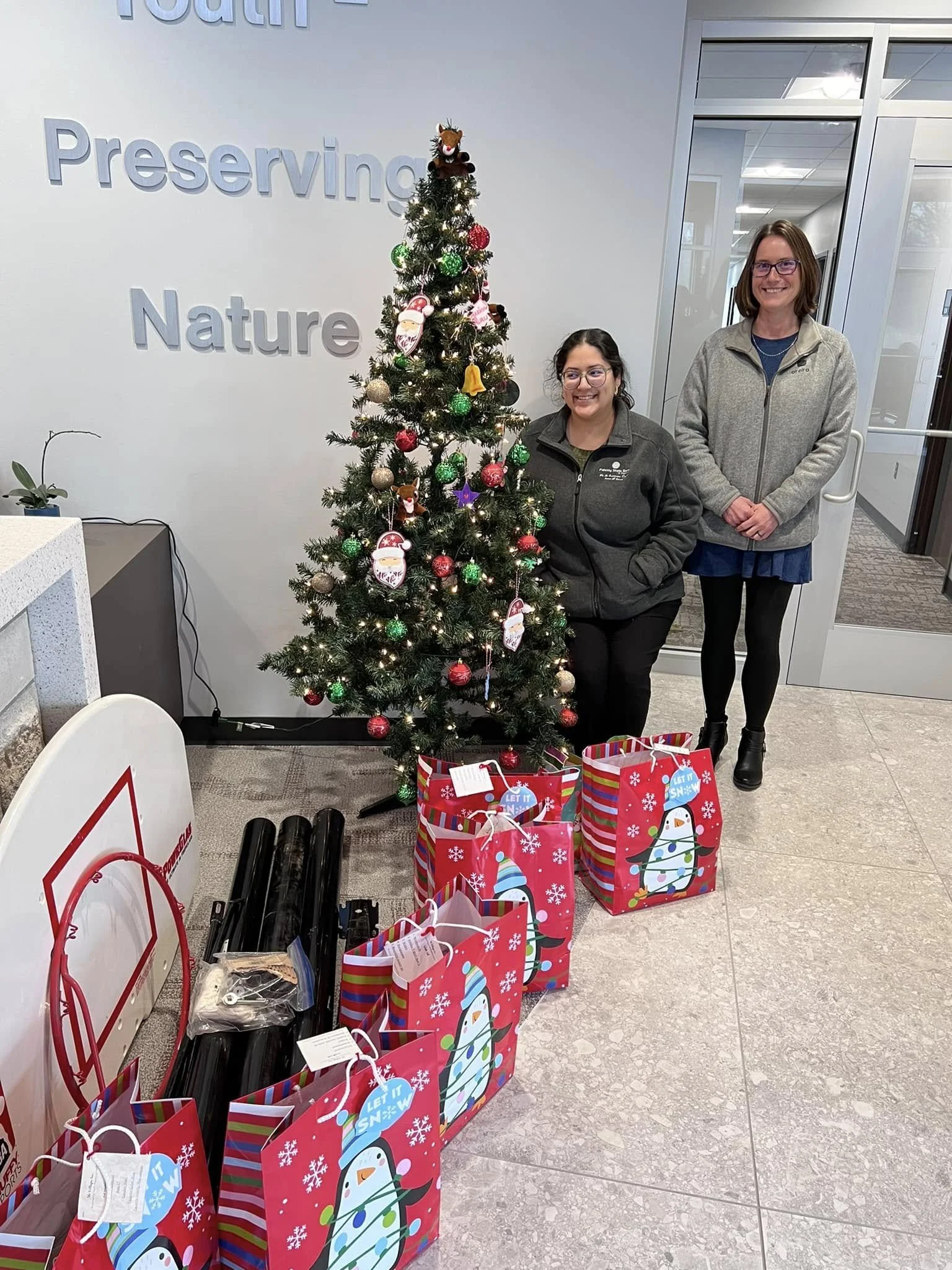 Two women standing next to a decorated Christmas tree with wrapped gifts underneath, inside an indoor space with a wall that has the word 'Preserving' and part of the word 'Nature' visible.