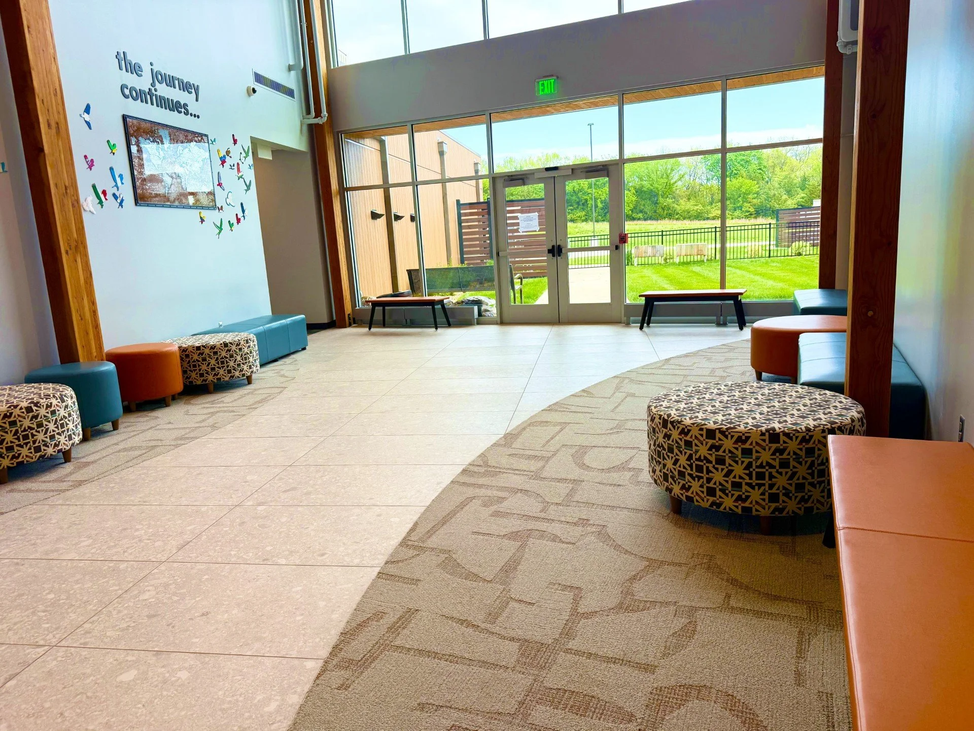 Indoor waiting area with large glass doors, colorful circular and rectangular seating, decorative wall with the phrase 'the journey continues...' and colorful bird decorations, overlooking a grassy area with trees.