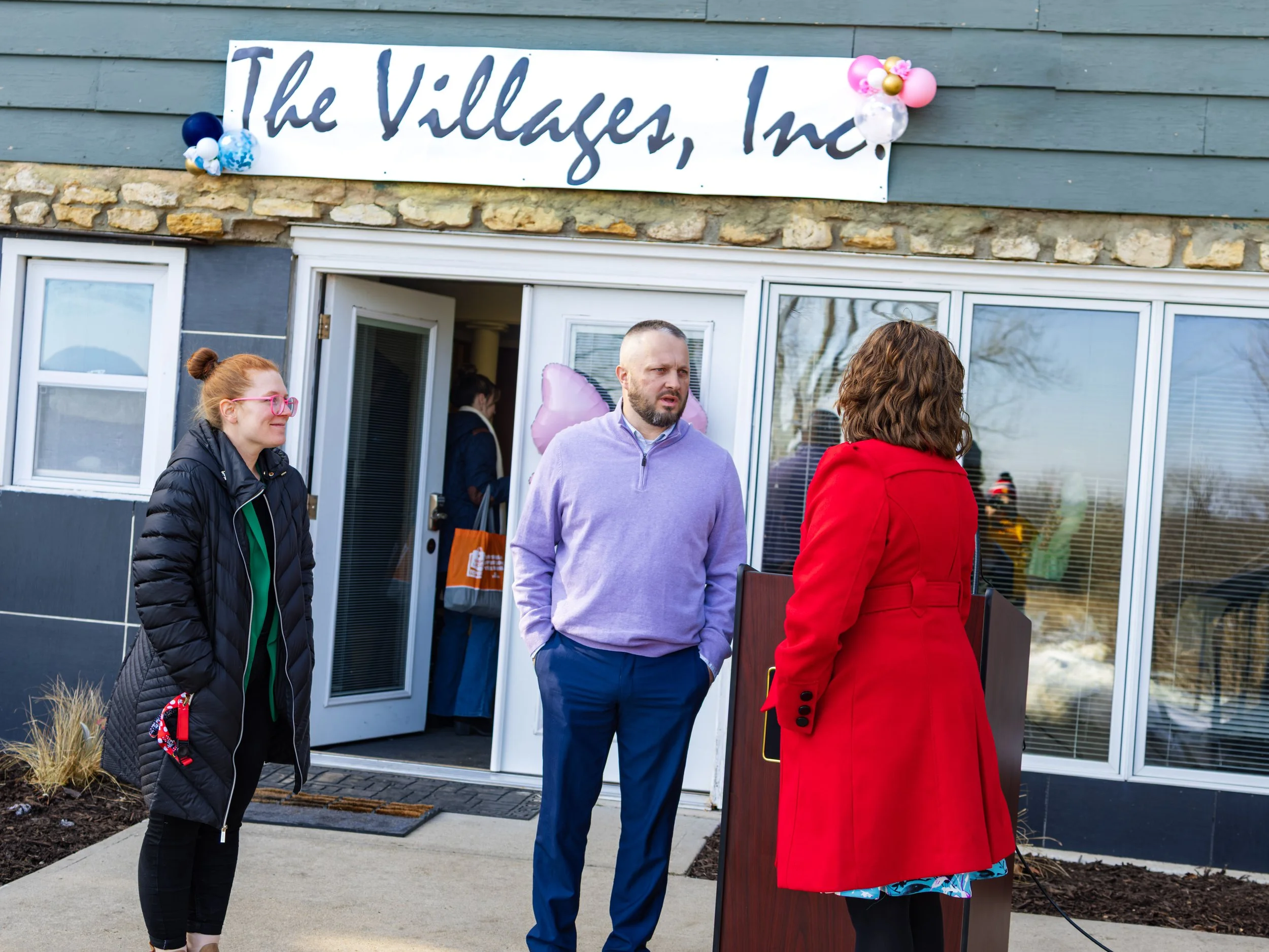 People standing outside in front of a building with a sign that says 'The Villages, Inc.' decorated with balloons.