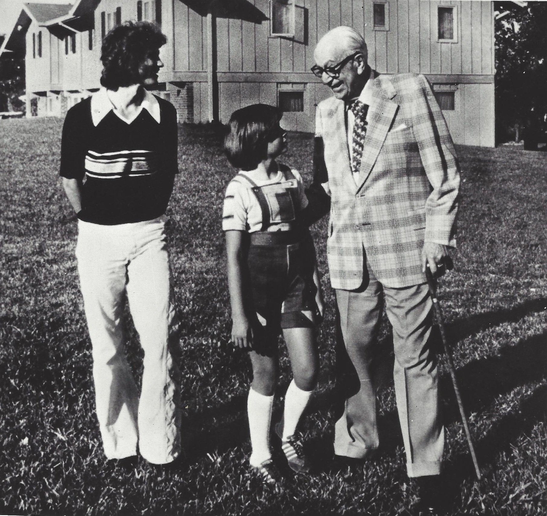 Four people standing outside near a building, engaged in conversation. Two adults, a woman and an older man, along with a young girl. The woman has curly hair and is wearing a sweater with a white collar, the girl has short hair and glasses, the man has gray hair, glasses, and is dressed in a plaid suit.