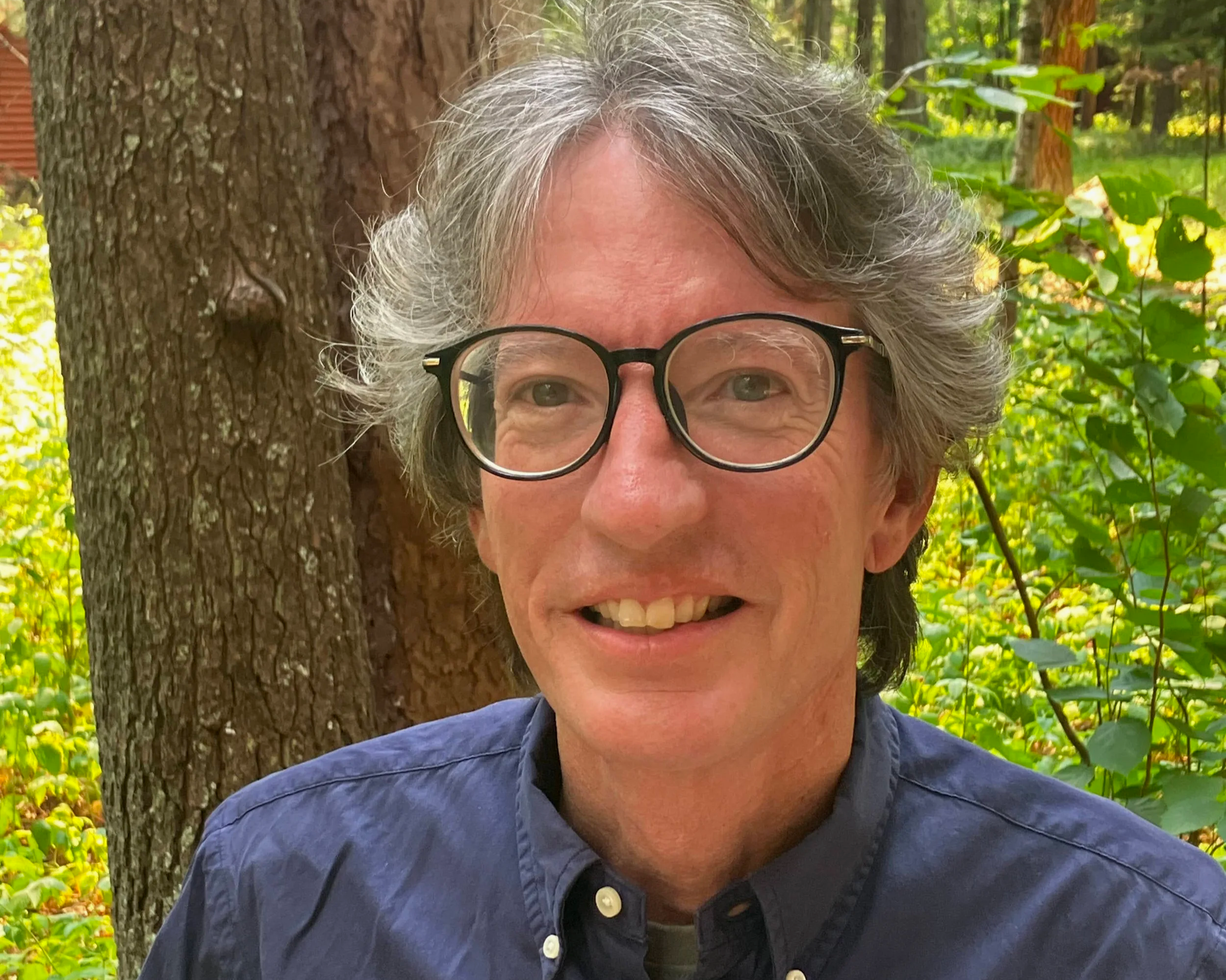 A woman with curly gray hair and glasses smiling outdoors near a tree, with a forested background.