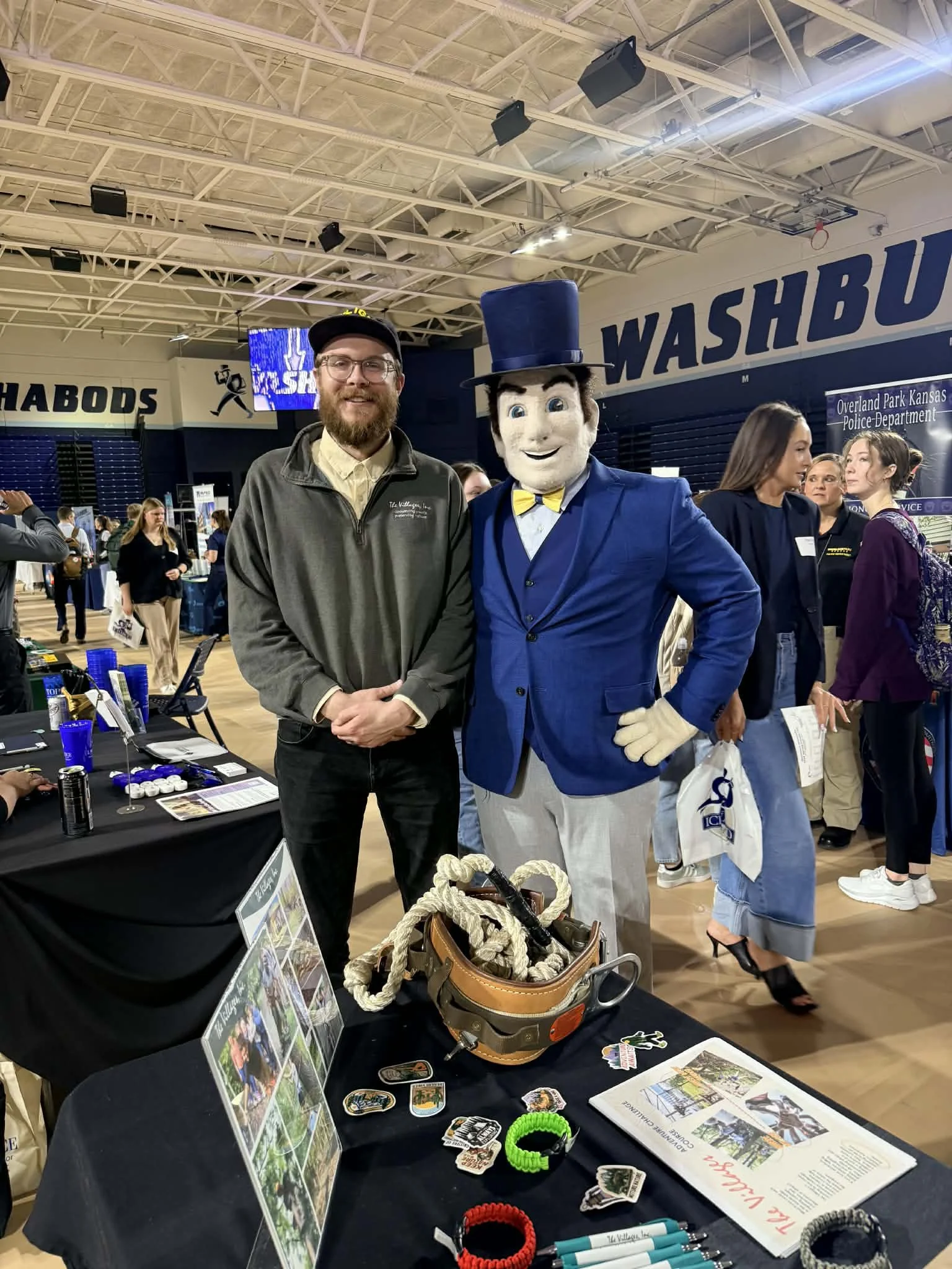 A man with glasses, a beard, and a black cap standing next to a person in a blue suit and top hat at an indoor event. There are multiple people in the background and a table with various items in the foreground.